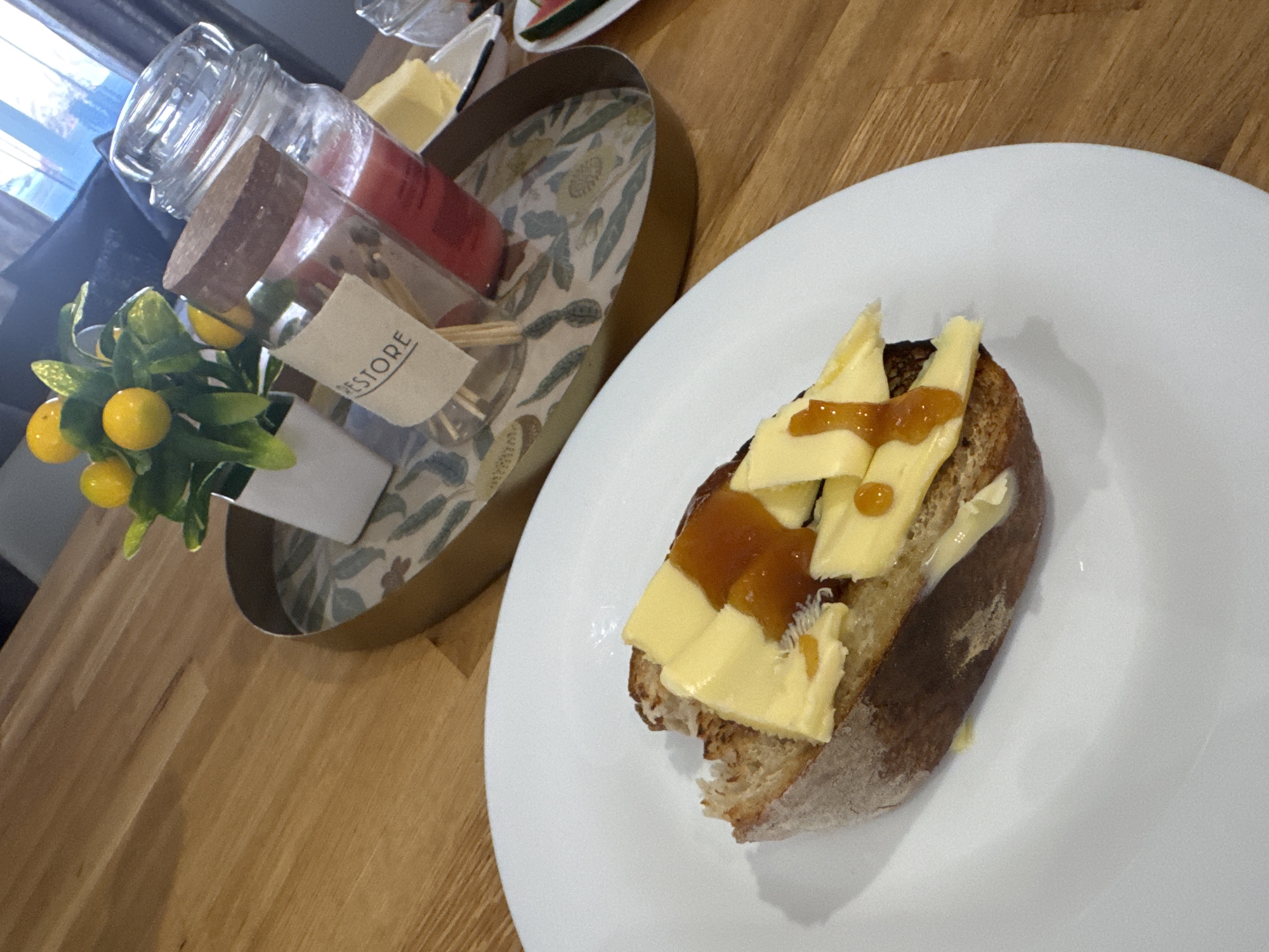 A slice of dark, crusty bread is topped with several thin slices of pale yellow butter and drizzled with a thick, amber-colored jam or preserves. The bread is served on a white plate, which is placed on a light brown wooden table. In the background, a gold-rimmed tray holds a small potted plant with bright yellow berries and green leaves, two glass jars with wooden lids, one containing red liquid and the other empty, and a piece of butter on a small dish. The scene appears to be indoors, possibly in a home environment in Dacorum, United Kingdom.