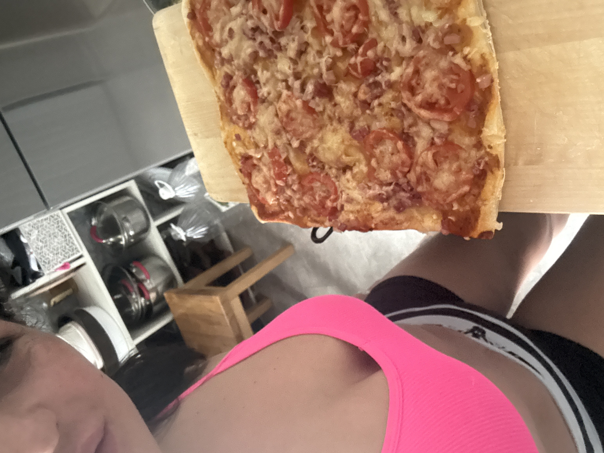 This image captures a close-up, angled view of a person with a rectangular pizza in an indoor setting, identified as Kauchuk Stantsiya, Russia.
The primary subject is a person, likely female, whose face is partially visible in the lower-left quadrant, showing part of the left eye, nose, and forehead. The person has dark hair and appears to have a neutral to slightly content expression. They are wearing a bright neon pink top, possibly a sports bra or tank top, and dark shorts with white stripes along the side.
The main object of focus is a freshly prepared, rectangular pizza resting on a light-colored wooden cutting board or pizza peel. The pizza has a thin, golden-brown crust and is generously topped with melted cheese, numerous slices of red tomatoes, and small, reddish pieces of what appears to be ham or pepperoni scattered across the surface.
In the background, to the left of the pizza, an interior space with storage shelves is visible. These shelves hold various household items, including several reflective metal pots or containers, clear plastic containers, and other items, some of which are wrapped in plastic. A small wooden stool or stand is also discernible in the mid-ground. The lighting suggests an indoor environment. There is no discernible text within the image. The scene conveys a casual, domestic moment, possibly related to cooking or enjoying a meal.