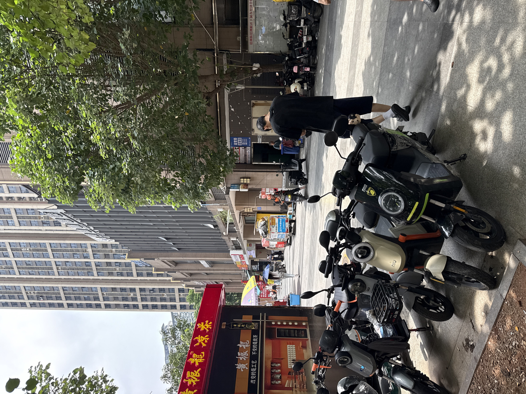 The image shows a street scene in Haizhu District, China. In the foreground, several electric scooters are parked along a sidewalk. A man in a black t-shirt and dark shorts stands with his back to the camera, looking at his phone. In the background, a tall, modern building with many windows dominates the skyline. A red awning with Chinese characters indicates a shop or business. Further down the street, a person walks with an umbrella, and a few delivery vehicles are visible. Trees with green foliage line the street, casting dappled shadows on the pavement. The overall impression is of a busy urban environment.