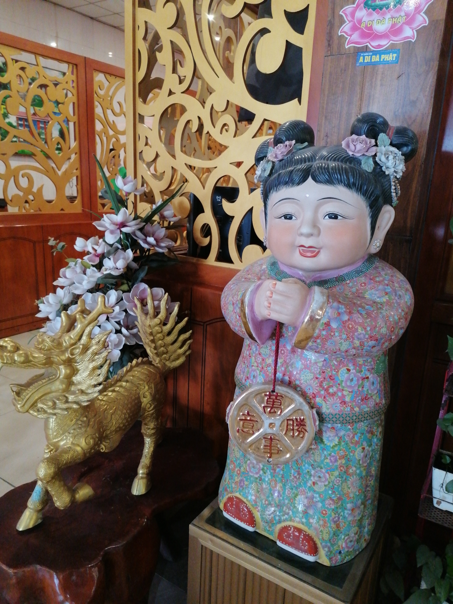 The image captures a scene in what appears to be an establishment in Chu Văn An Ward, Vietnam.
To the left, a golden dragon statue sits on a dark, intricately carved wooden stand. Adjacent to the dragon is an arrangement of white and pale pink flowers.
To the right of the dragon, a statue of a child-like figure dressed in a colorful, patterned garment stands on a small wooden pedestal. The figure has two buns with flowers. A round coin hangs from the figure's hands.
The background features wooden decor and a sign at the top with "A Di Đà Phật" on it.