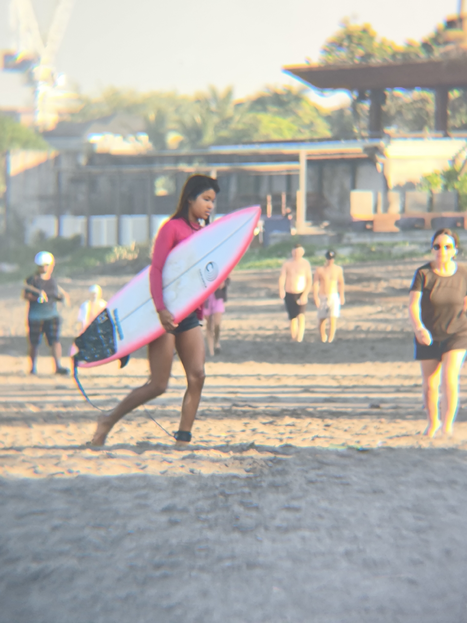 The image depicts a beach scene in North Kuta, Indonesia. A young woman is carrying a surfboard with a pink outline on the beach. She is wearing a long-sleeved pink shirt and dark shorts. The surfboard has a leash attached to her ankle. Several other people are visible in the background, including two men in swimwear, one person in a light-colored shirt and dark shorts, and a woman in a dark shirt and shorts. The backdrop shows a building and trees. The beach sand is light-colored, and the overall lighting suggests it is daytime.