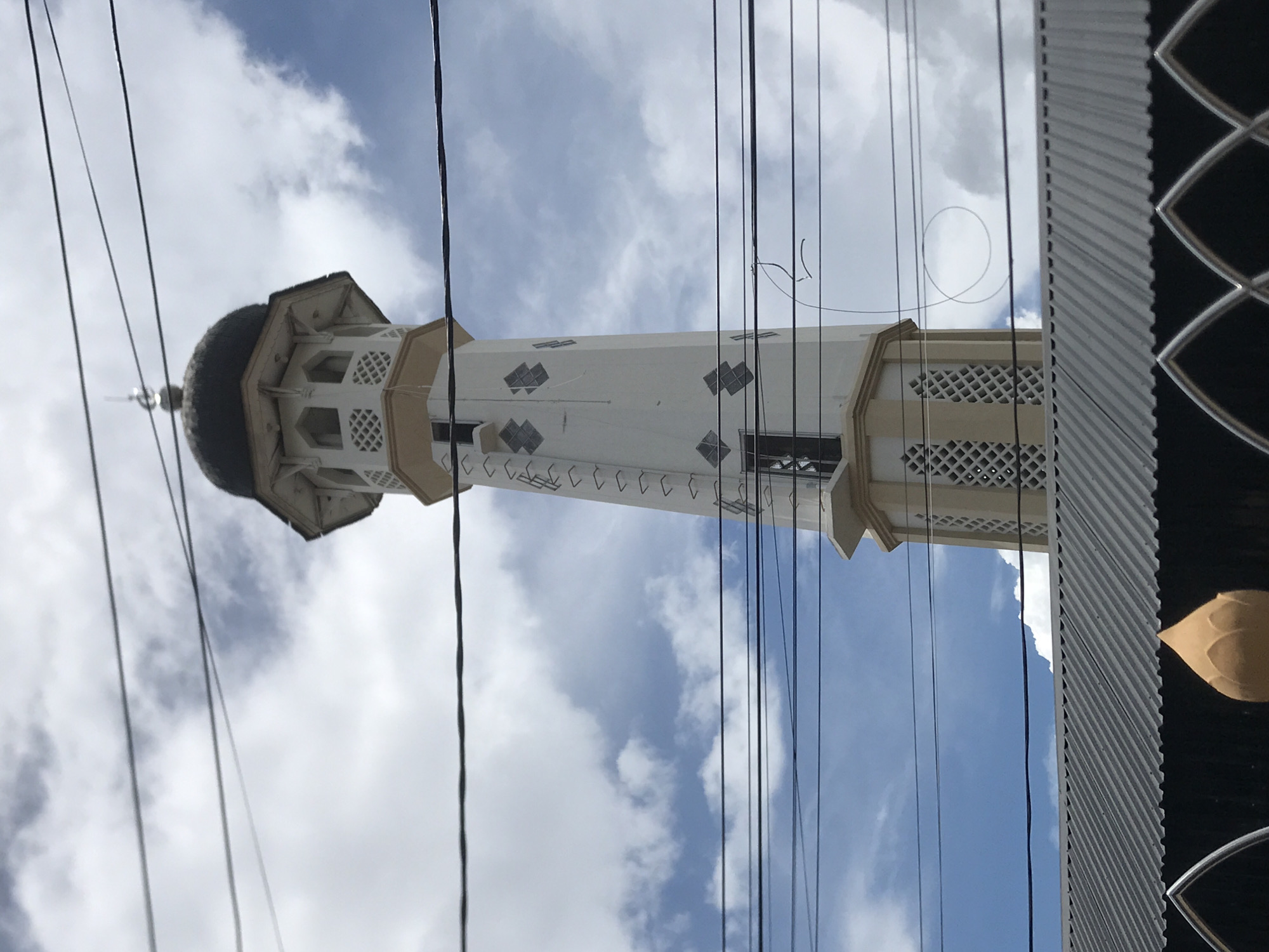 The image shows the minaret of a mosque in Lhokseumawe, Indonesia. The minaret is tall and slender, with a black dome at the top. It is decorated with geometric patterns and has small windows or openings along its length. The minaret is white with tan accents. Power lines run across the foreground. The sky is visible in the background, with white clouds and patches of blue. Part of a building with decorative features is visible on the right side of the image.