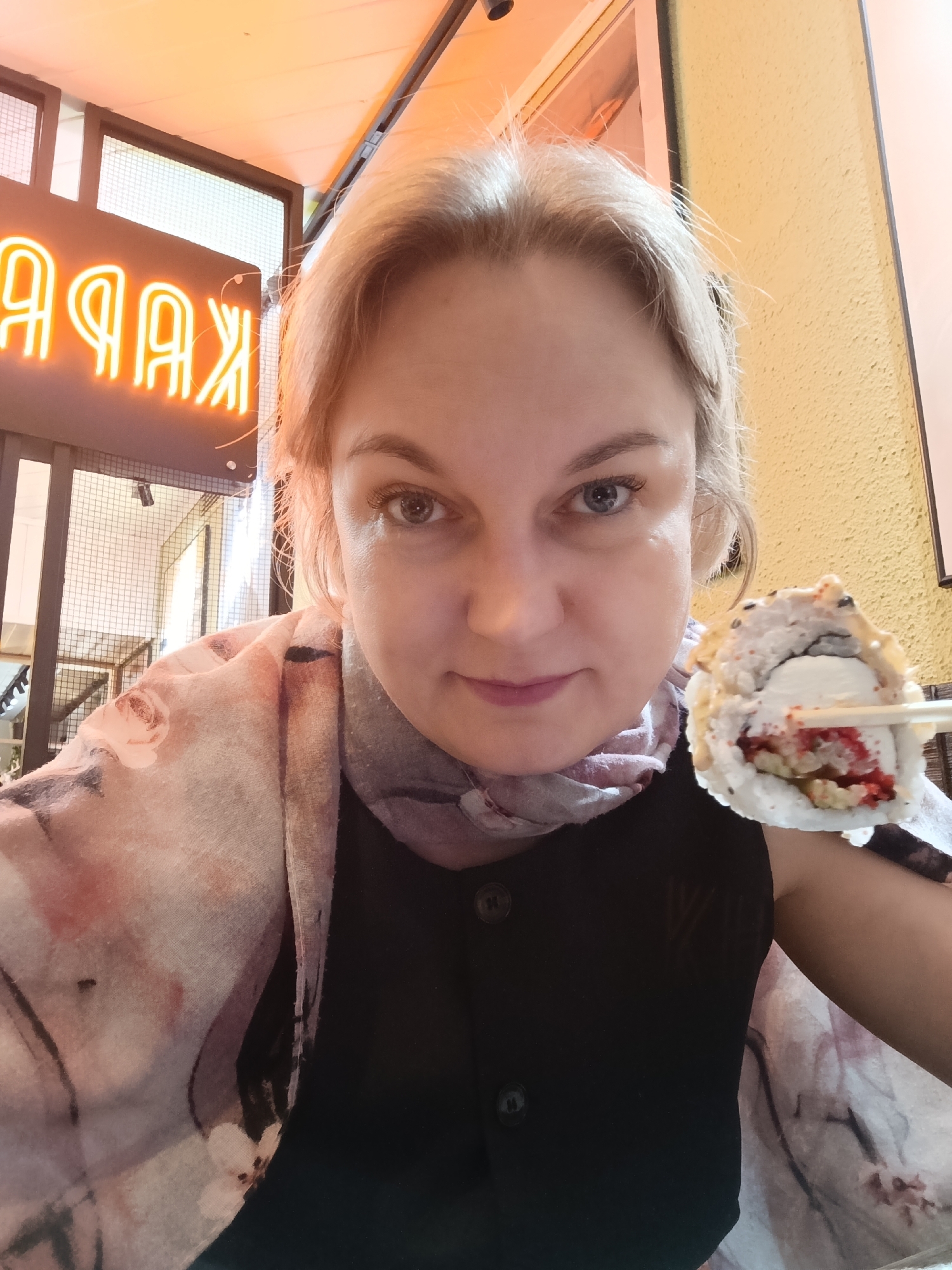 A woman with fair skin and light blonde hair takes a selfie in what appears to be an indoor dining establishment in Chaikovsky, Russia. She looks directly at the camera with a neutral expression. She is wearing a dark, possibly sleeveless top with two visible buttons on the front, and a light-colored scarf with a soft floral pattern in pinks, purples, and grays draped over her left shoulder and neck.
In her right hand, held by a pair of light-colored chopsticks, she presents a single piece of sushi. The sushi roll is distinctive, featuring white rice, a generous portion of a creamy white filling (possibly cream cheese or a similar ingredient), and bright red roe or caviar. The exterior of the roll is lightly coated with small dark specks, possibly sesame seeds.
The background is softly out of focus, indicating an indoor setting. To the left, a bright orange neon sign is prominent, displaying Cyrillic letters that appear reversed, reading "ЯАРАК" (likely "KAPAA" or "KAPAK" when correctly oriented). Below the sign, a dark-framed structure with a grid pattern is visible, possibly a window or a decorative screen. To the right, a textured yellow wall forms part of the interior decor. The overall lighting in the image is bright and warm.