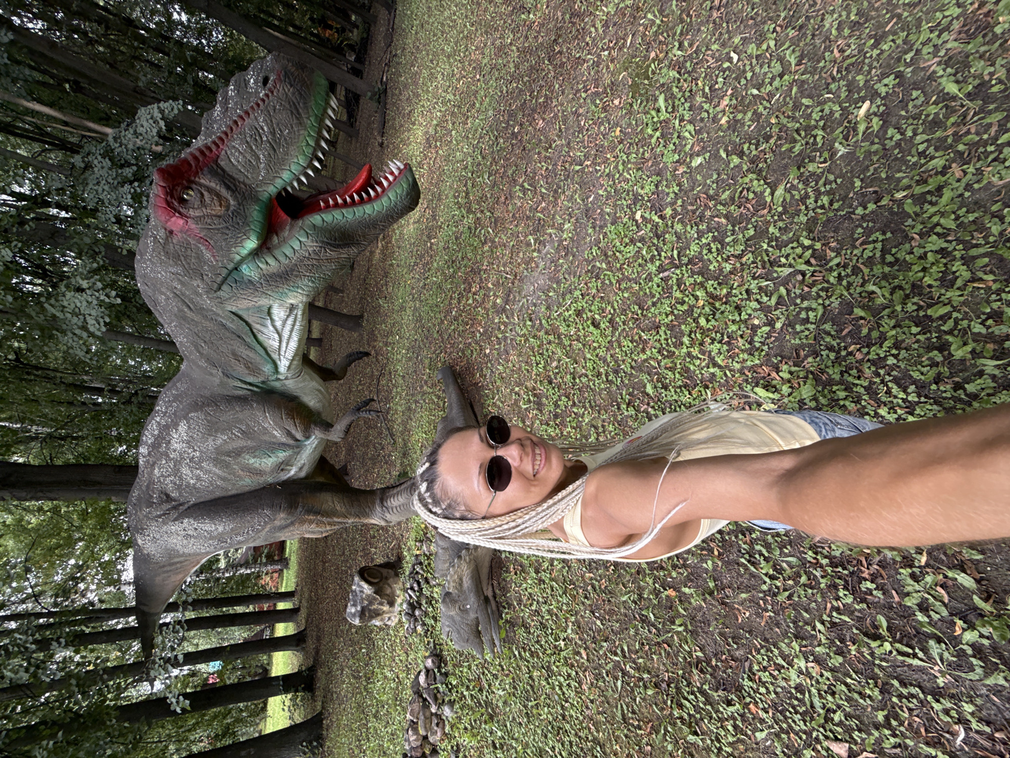 A woman with blonde braids and sunglasses smiles at the camera while taking a selfie. She is wearing a yellow tank top and blue jeans. Behind her, a large dinosaur statue with its mouth open is on display in a wooded area. Another dinosaur statue is lying on the ground in front of it. The ground is covered in grass and fallen leaves.
