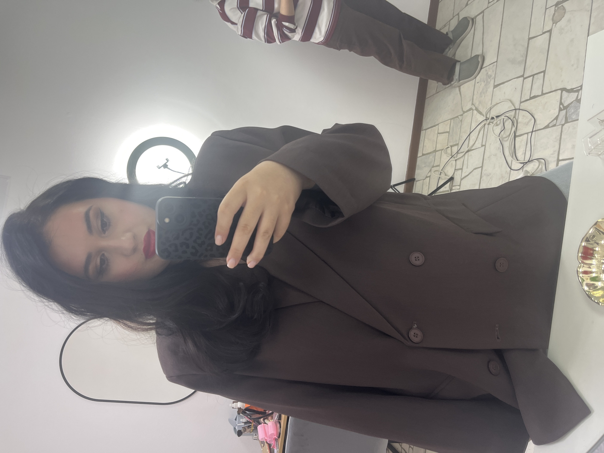 A young woman takes a selfie in an indoor environment. She is wearing a brown blazer and has dark hair and red lipstick. A ring light is behind her. Another person wearing a striped shirt and brown pants is partially visible in the background. The floor is tiled. It's difficult to confirm the location as Chaikovsky, Russia, as there are no specific geographical cues.