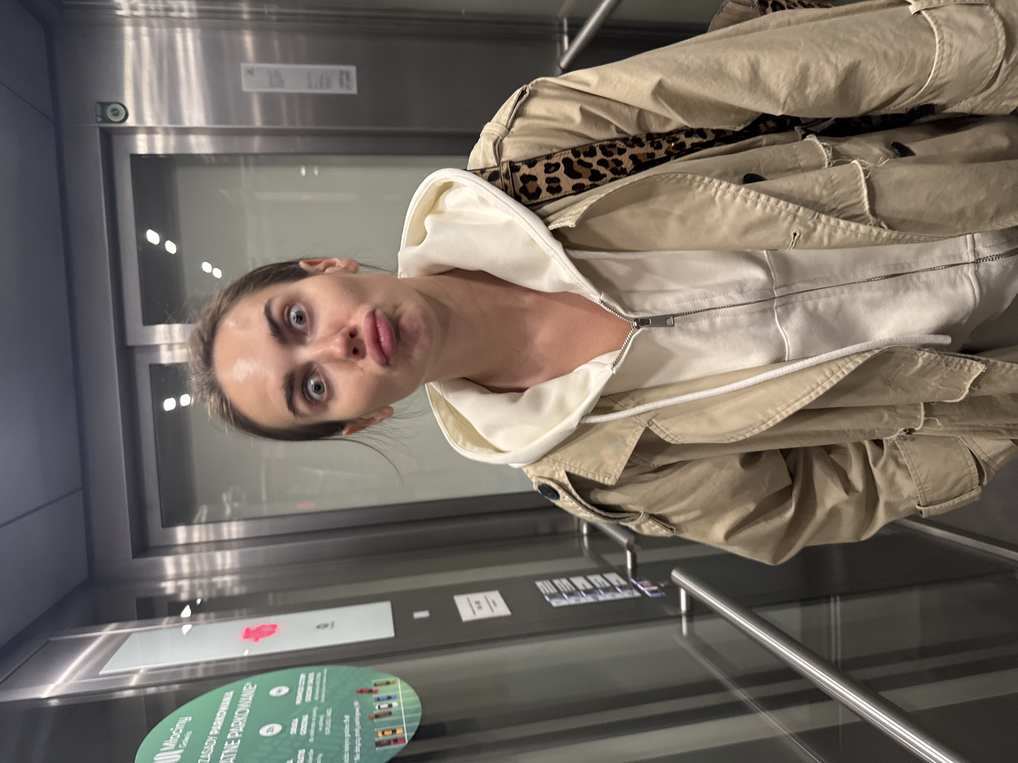 The image captures a young adult woman making a humorous, exaggerated expression within what appears to be a modern elevator or building interior in Warsaw, Poland. The woman, positioned prominently in the frame, looks directly at the viewer with wide eyes and puckered lips. She has dark hair and is wearing a light-colored, possibly off-white, hooded sweatshirt beneath a beige or khaki trench coat; a leopard print strap or lining is visible over her shoulder.
The setting features sleek, metallic silver walls and large, reflective glass panels, creating multiple reflections of the environment and the subject. To her right, a control panel with numerous small, square buttons indicates an elevator. A metallic handrail extends horizontally across the lower portion of the scene.
On the left side of the image, behind a glass panel, two signs are visible. The most prominent is a circular, green and white sign with colorful car illustrations and Polish text. Key phrases on this sign include "Młociny" (a district in Warsaw), "ZASADY PARKOWANIA" (Parking Rules), and "KREATYWNE PARKOWANIE" (Creative Parking), along with words like "OGRODZIEC," "STREFA," "GODZINA," and "OPŁATY." Below this, a rectangular sign with a red icon is also present. The reflections on the glass surfaces show blurred elements of the surroundings and bright overhead lights. The overall mood is playful and candid, captured in an urban, contemporary indoor environment.