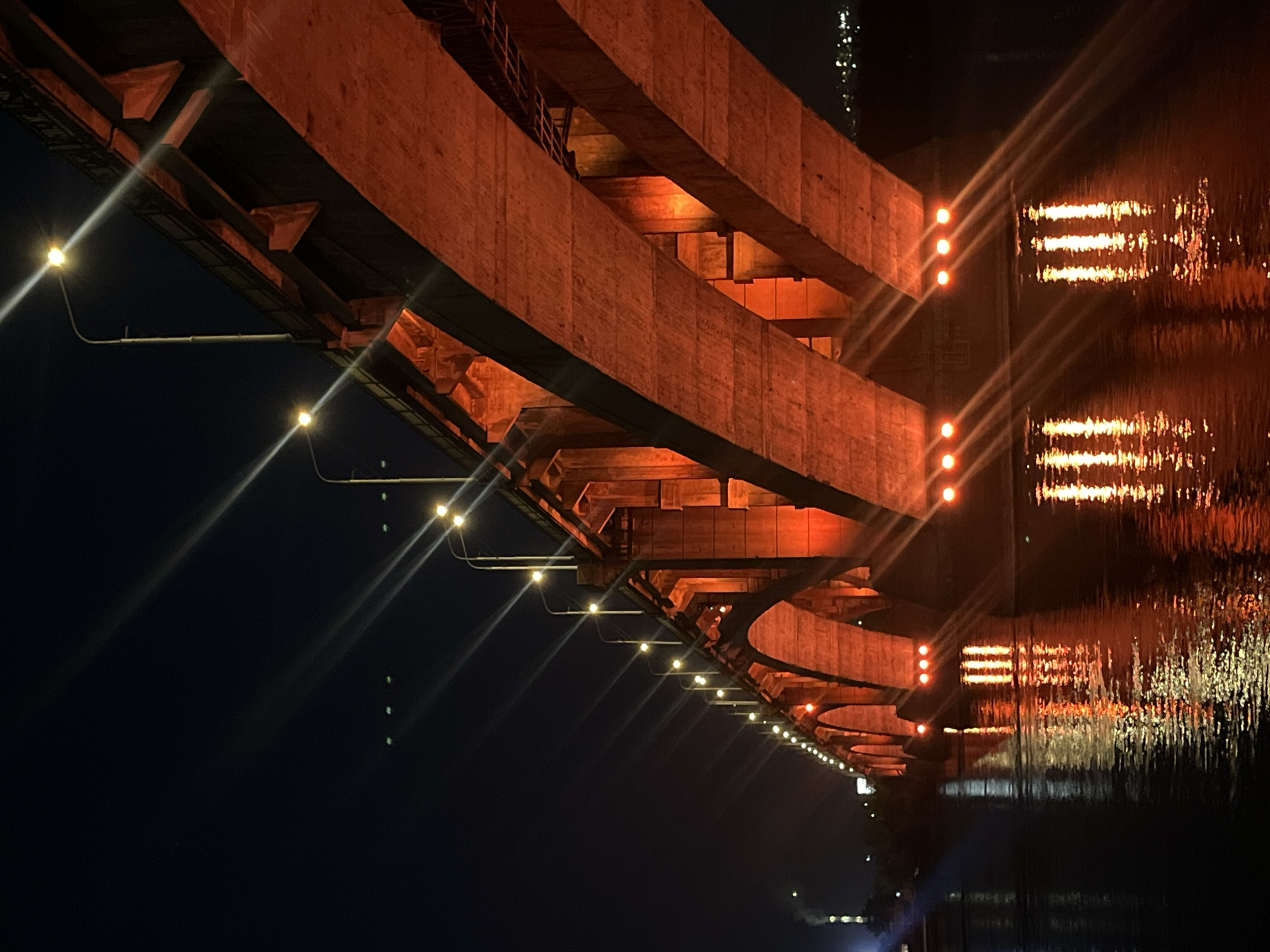The image presents a nighttime, low-angle view of the underside of a large bridge, likely spanning a river or a significant body of water, evidenced by the reflective surface on the right. The scene is devoid of people or discernible activity, focusing instead on the bridge's architectural structure and its artificial illumination.
The bridge features robust construction, with thick concrete or metal beams, girders, and supporting structures visible from below. These elements are bathed in a warm, intense orange-red light, suggesting upward-facing spotlights that highlight the bridge's impressive scale and material texture. Along the lower edge of the bridge's span, a series of smaller, yellowish-white lights are installed in a linear sequence, providing direct illumination. These lights create prominent starburst effects or lens flares, streaking across the dark, clear night sky.
The water on the right side of the frame acts as a dynamic mirror, reflecting the bridge's lights. The orange structural lighting appears as broad, horizontal bands of light on the rippled water surface, while the smaller yellowish lights are reflected as elongated, distorted streaks. The overall lighting scheme emphasizes the industrial grandeur of the structure against the deep black of the night. The time of day is clearly night, with no indications of specific weather conditions other than a calm surface for reflections. There is no visible text in the image. The location is Krasnoyarsk, Russia.