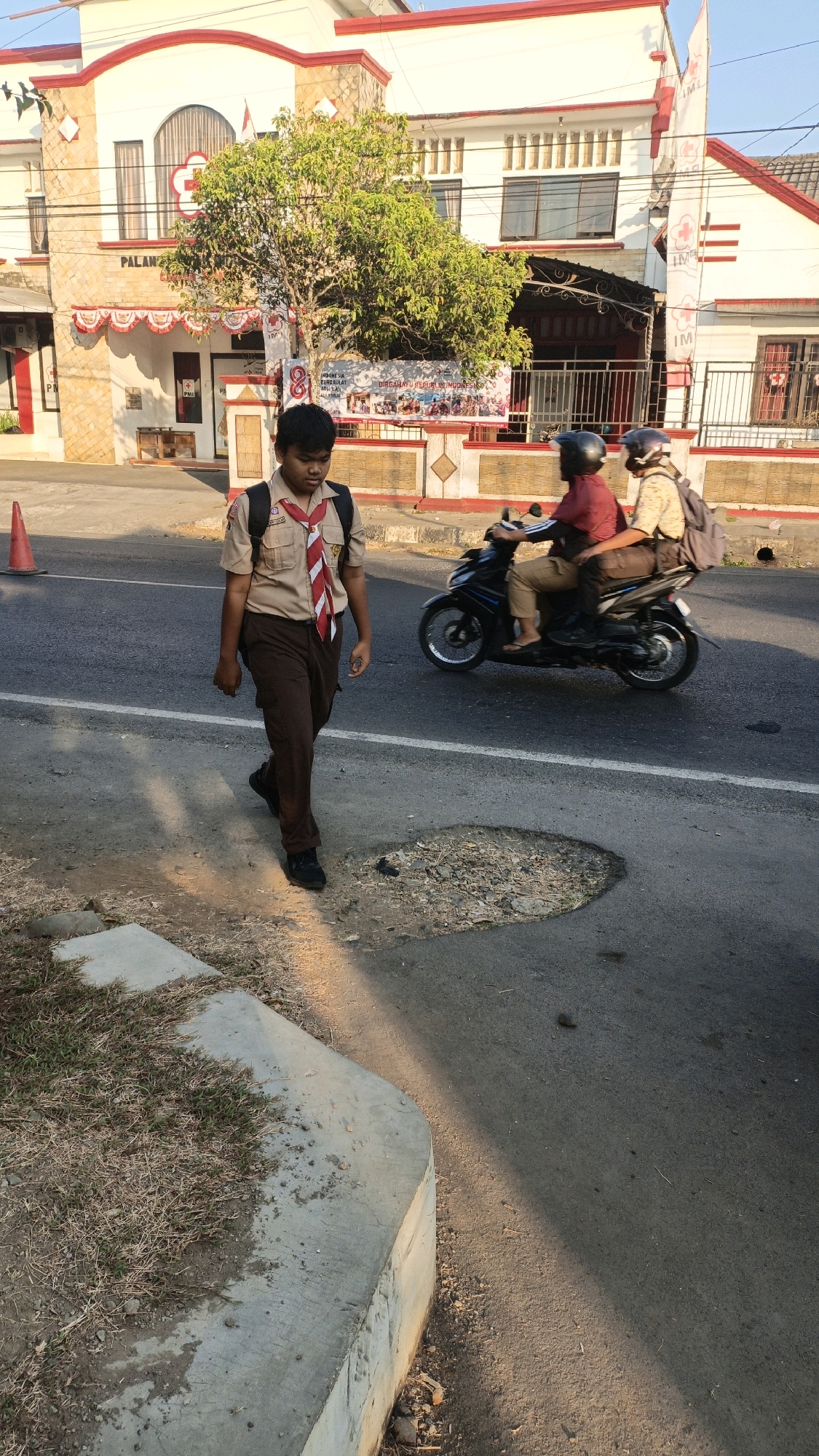 In Kuwasen, Indonesia, a young boy wearing a scout uniform walks on the side of a road, his backpack slung over his shoulders. He is on the left side of the frame, and the road appears to be in a town or city, with a building in the background. The building is white with red trim and appears to be a public building, possibly a clinic or government office, with the letters "PMI" visible. A tree with lush green leaves stands in front of the building. On the right side of the road, two people are riding a scooter. The scooter is black, and the riders are wearing helmets. The road itself has a large pothole. The sky is clear and the lighting suggests it is daytime. The overall scene depicts a common day in a populated area of Indonesia.