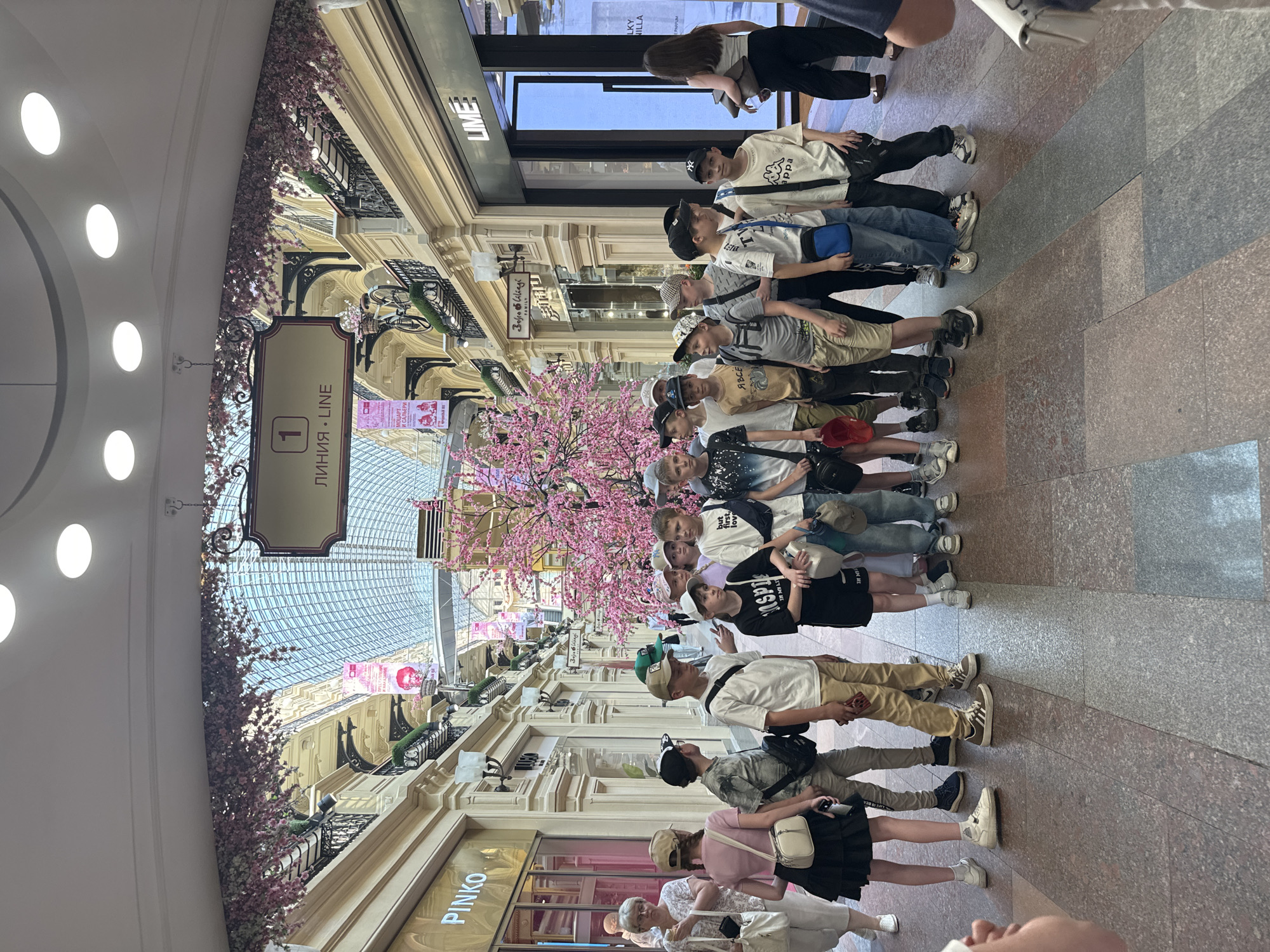 A crowd of around 15 people, mostly young boys, is standing in the main hall of GUM, a large department store in Moscow, Russia. The hall is decorated with pink cherry blossoms, and shops line the sides of the walkway. The boys are wearing casual clothing, and some are carrying bags. Some adults are standing in the background. The hall has a glass ceiling, and a sign indicates "Line 1" in Russian and English. Several signs related to shops are visible.