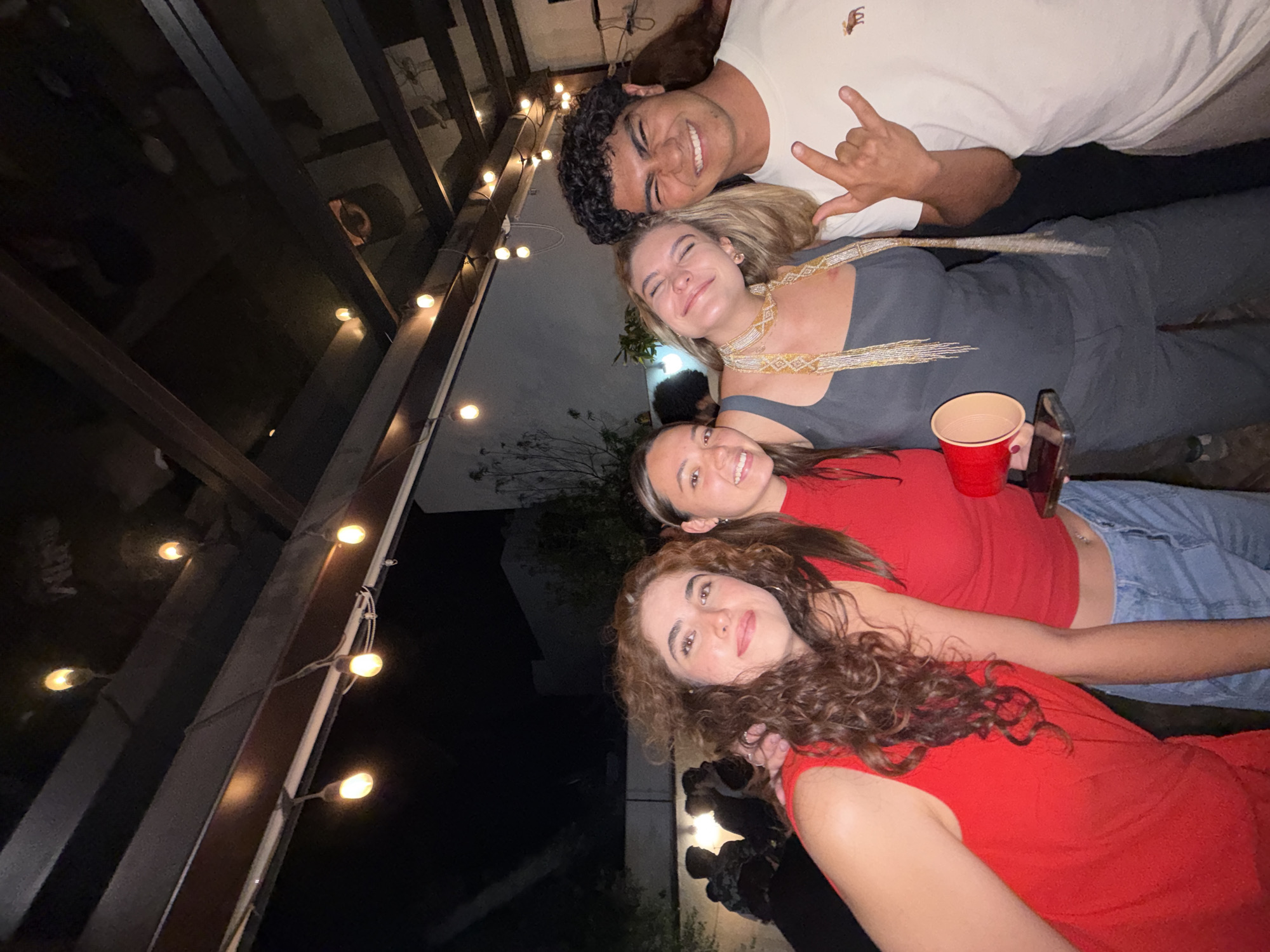 The image shows a group of four people posing for a photo in an outdoor setting at night. The man at the top is smiling and making the "rock on" hand gesture. Below him is a woman in a gray outfit with a gold beaded necklace. Underneath her is another woman in a red top holding a red plastic cup and a phone. At the bottom is a woman with curly hair in a red top. String lights are visible in the background, suggesting a patio or outdoor gathering. The location is Unknown, Unknown.