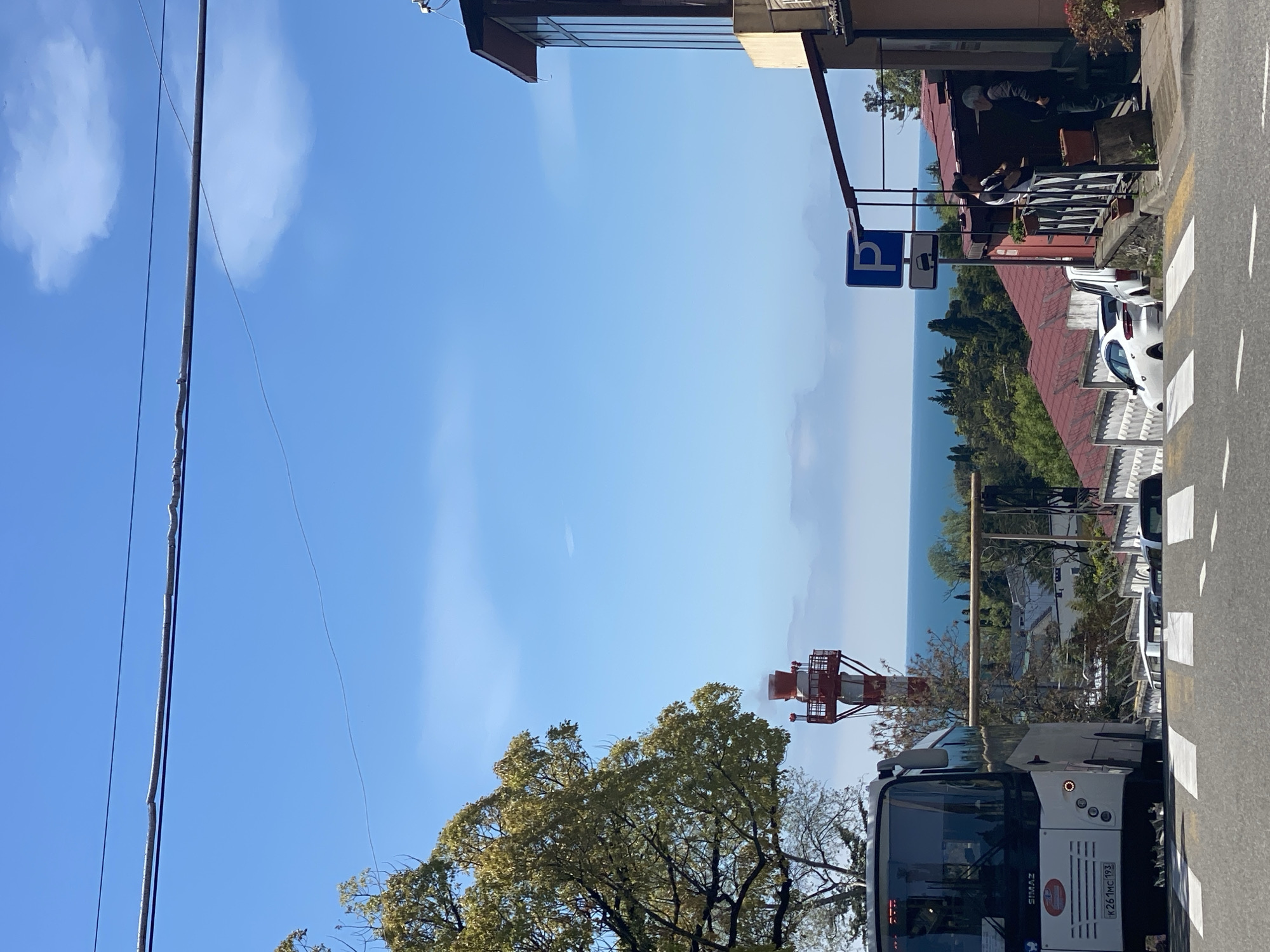 The image presents an outdoor scene with a clear blue sky and scattered clouds. A street is visible on the right side of the frame with a white bus and parked cars. On the left side, power lines intersect the sky. A building with a balcony is situated on the right, and a parking sign is visible. In the middle ground, a red and white communication tower rises above the treeline. The Black Sea can be seen in the background. The bus registration plate reads "K26 MC 193", indicating a vehicle registered in Russia. The location is Sochi, Russia.