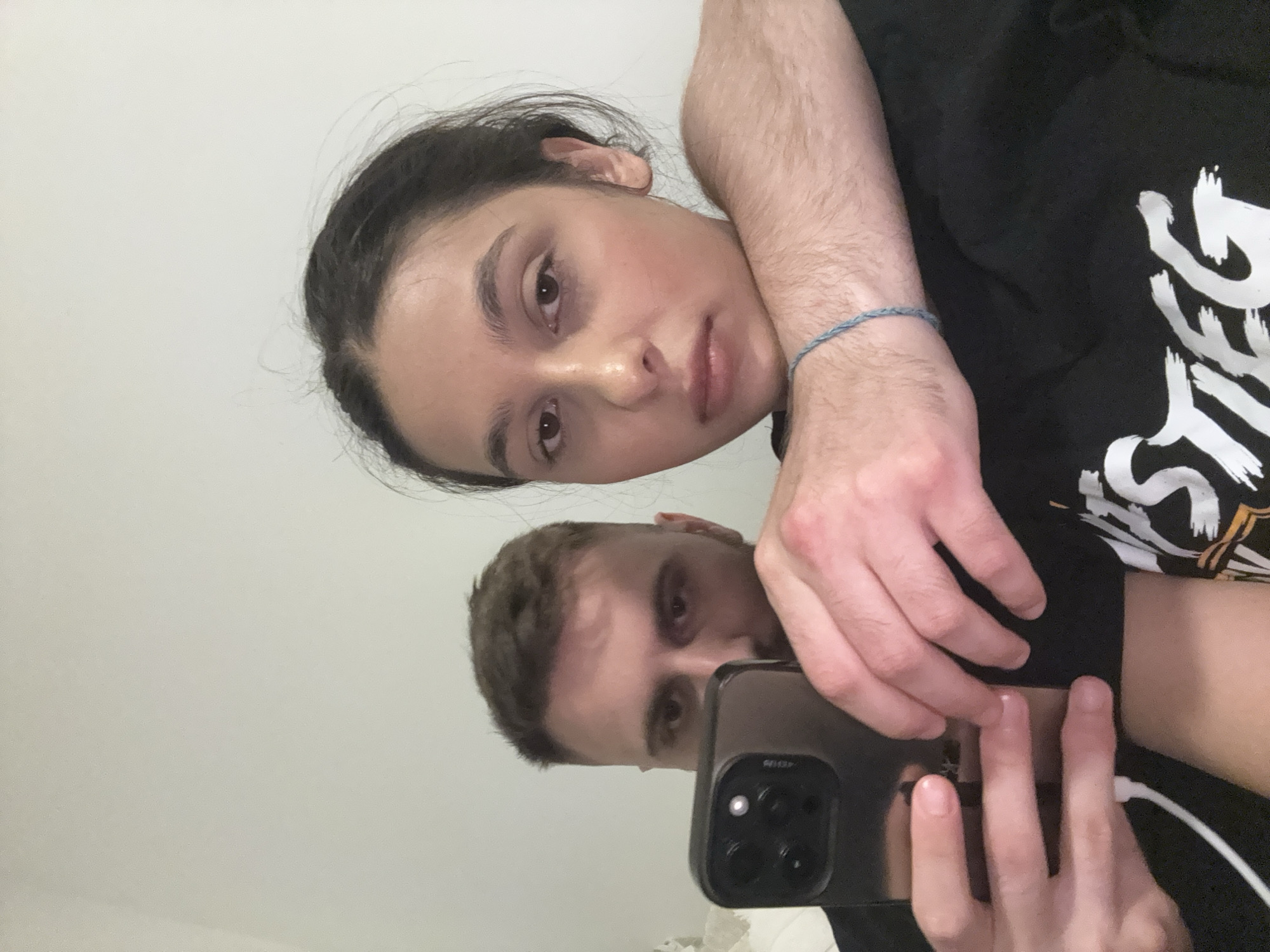 The image is a close-up selfie taken by two individuals, a man and a woman, with a plain white wall in the background. The man is on the left, holding a smartphone with a dark case, and is partially visible, looking towards the phone. The woman is on the right, her face is the primary focus, and she appears to be leaning on the man's arm. The man's arm, with visible hair, is wrapped around the woman's shoulder and chest area. The woman is wearing a black t-shirt with white lettering, part of which reads "STEG". A thin blue string bracelet is visible on the man's wrist. The lighting suggests an indoor setting, possibly a bedroom or living area. The image does not contain any identifiable landmarks or features that would place it specifically in Mönchengladbach, Germany.