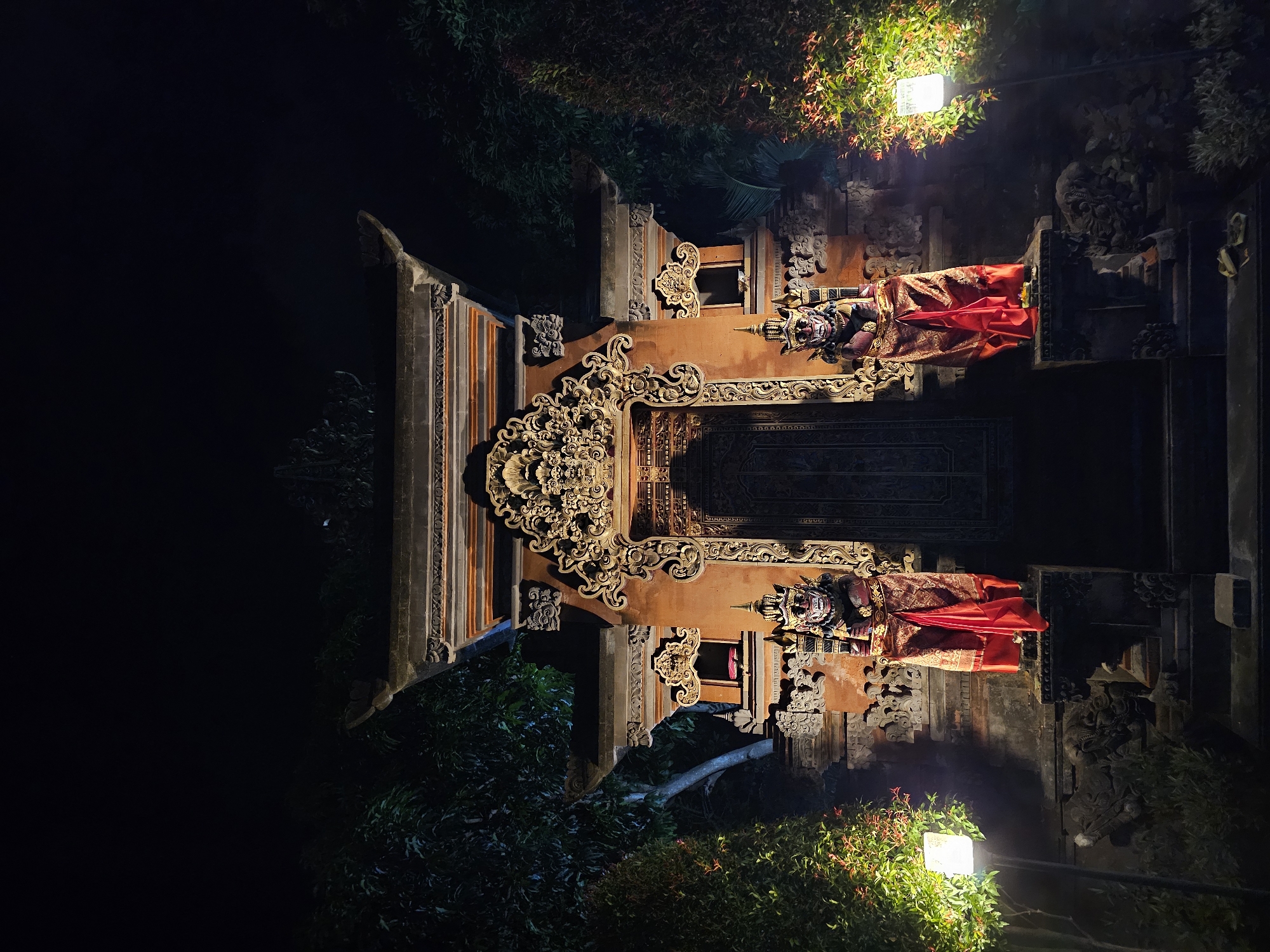 This is a nighttime, vertical, eye-level shot of an ornately decorated doorway, likely part of a temple or traditional Balinese building. The doorway is a deep reddish-orange color and is flanked by two elaborate statues. Each statue is adorned with a richly patterned red and gold fabric, suggesting a ceremonial or festive occasion. Above the statues, intricate stone carvings are visible, featuring floral and mythical motifs. The doorway itself is also heavily carved, with a dark, textured surface.
The scene is illuminated by two bright, square lights, one positioned above and to the right of the doorway, casting a warm glow on the foliage and upper portion of the structure. Another light is visible lower down on the right side, also illuminating plants. Dark, lush green foliage surrounds the structure, creating a stark contrast with the illuminated architecture. The background is almost entirely black, indicating it is deep night.
The orientation of the image is vertical, with the doorway appearing to be in the center of the frame. The overall mood is one of mystery and grandeur, with the vibrant colors of the fabrics and the detailed carvings standing out against the darkness. The presence of the draped fabrics suggests that an event or ritual may have recently taken place or is about to commence. The location cues, such as the architectural style and the vegetation, are consistent with Denpasar, Indonesia. No visible text is present in the image.