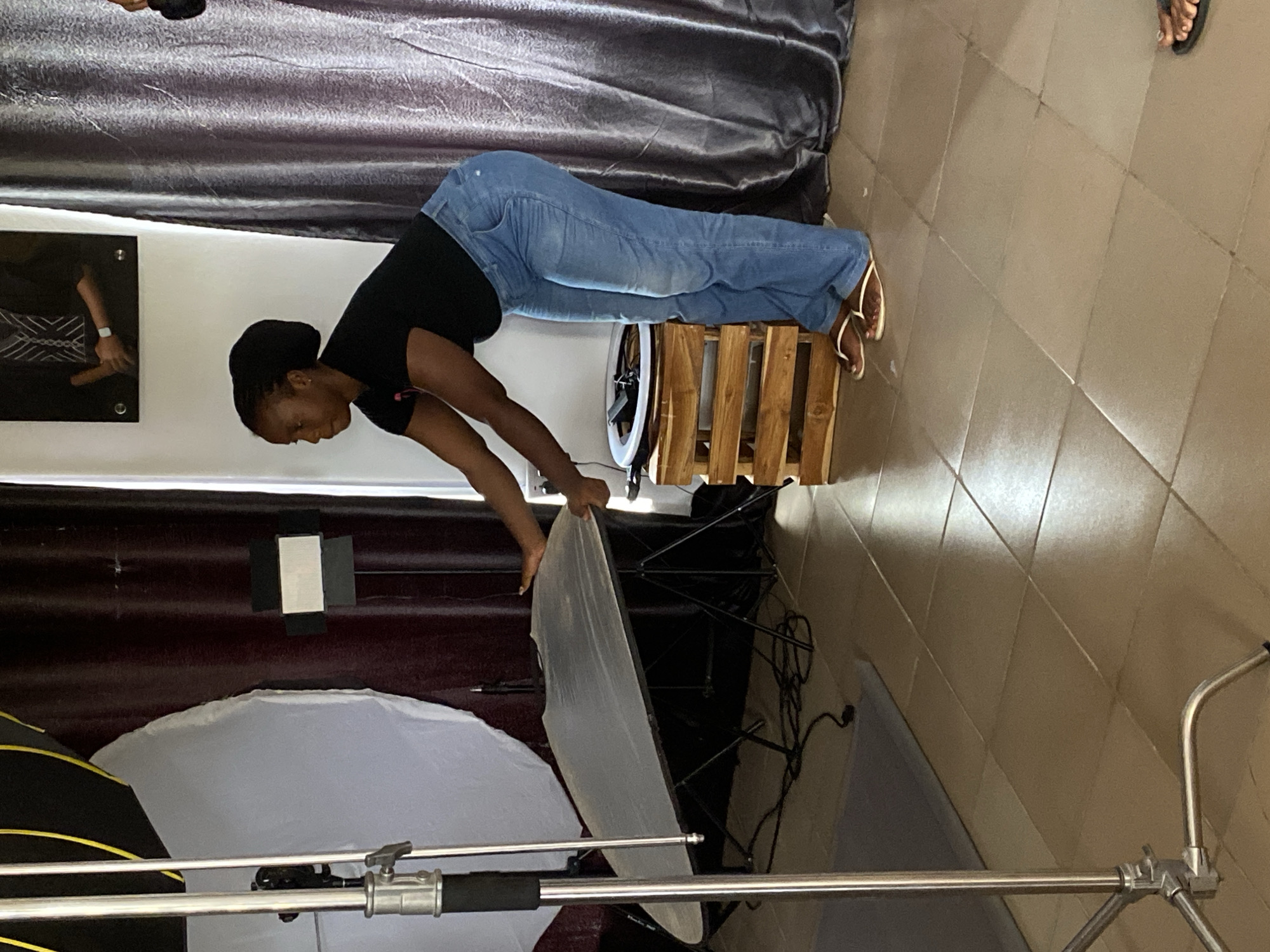 An indoor scene in Jos, Nigeria, depicts a woman in the process of setting up or adjusting photography equipment. The woman, with dark skin and hair tied back, is wearing a black t-shirt and blue jeans, along with light-colored flip-flops. She is leaning forward, holding a large, rectangular, white photography reflector or diffusion panel with both hands, positioned over a light stand.
The room features a light brown tiled floor and white walls. A dark, textured curtain or drapery covers the wall behind the woman and extends across the upper part of the frame. To her right, a light-colored wooden crate is visible, and behind it, a circular ring light is partially seen. On the left side of the image, a partially collapsed black softbox with yellow accents is visible on another stand. A black framed picture or canvas, displaying a portion of an arm wearing a watch and a white design, hangs on the white wall. The overall impression is of a practical photography workspace being prepared or utilized. The time of day is not discernible, as the setting appears to be lit by artificial light.