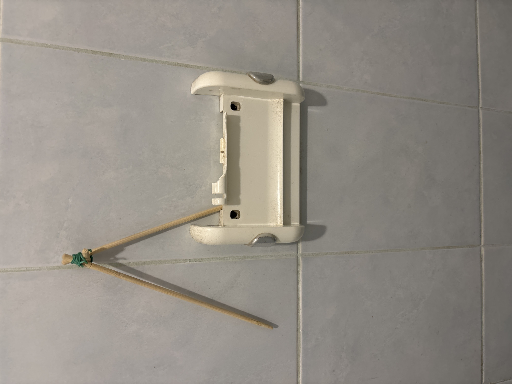 Two wooden chopsticks, bound together at the top with green twine, are suspended from a tiled wall, angled downwards and to the right. Below the chopsticks, a cream-colored plastic object, appearing to be a broken toilet paper dispenser, is mounted on the wall. The dispenser is chipped and shows signs of wear. The wall is covered in light gray square tiles with visible grout lines.