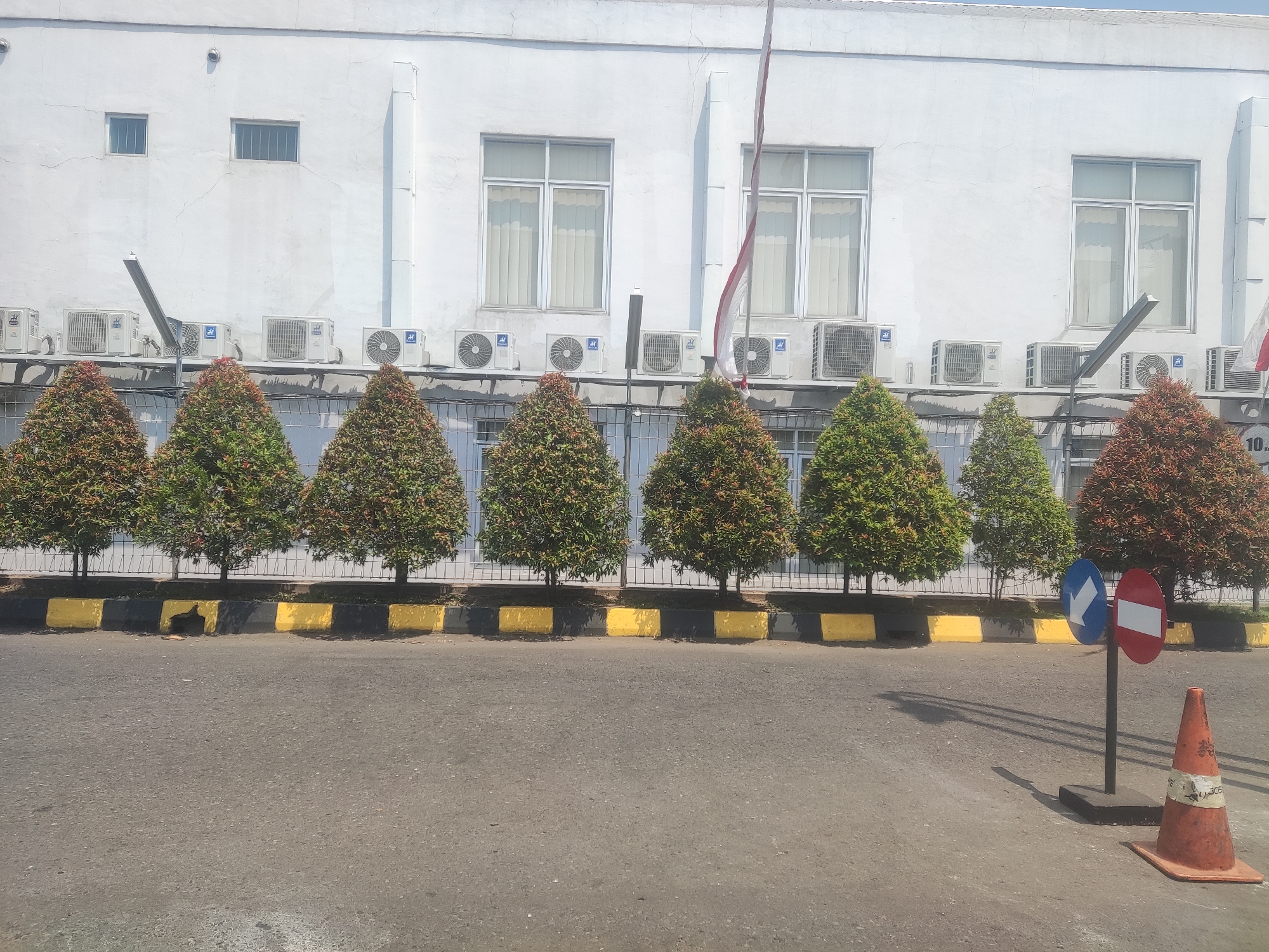 The image captures an outdoor scene in front of a white building. The building has multiple windows with blinds and several air conditioning units mounted on its exterior wall, below the windows. In front of the building, a row of neatly trimmed, conical-shaped trees with reddish-green foliage is planted along a curb. The curb is painted with alternating yellow and black blocks. Beyond the trees, a metal fence is visible. The ground in front of the curb is an asphalt road. On the right side of the road, near the curb, there are two traffic signs, one displaying a blue arrow pointing left and a white background, and the other a red and white circle with a horizontal bar. An orange traffic cone with white tape is placed on the road next to the signs. The sky is bright and appears to be daytime. The scene is devoid of people and any apparent activity. The location is identified as Trihanggo, Indonesia.