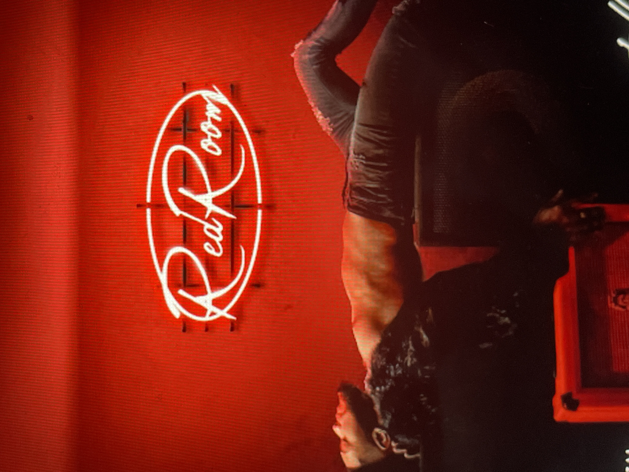 An indoor scene, likely in Sofia, Bulgaria, features a striking red aesthetic with a person performing an acrobatic inversion against a vibrant red wall. The image shows the person hanging upside down, dressed in dark, form-fitting attire. Their head is visible at the bottom of the frame, featuring dark, curly hair, and their left arm and upper torso are prominent as they hold the pose.
On the bright red wall behind them, a prominent white neon sign glows, clearly spelling out "Red Room" in a stylized, cursive font, enclosed within an oval shape. Horizontal white lines extend from either side of the oval. To the right of the person, partially obscured in shadow, is a dark area with a glimpse of an orange or red structure, possibly a prop or stage equipment.
The setting appears to be a performance venue or club, characterized by dramatic artificial lighting and a dominant red color scheme. The activity suggests a dynamic performance or artistic shoot. The notable details include the vivid red background, the glowing "Red Room" sign, and the person's athletic pose, all contributing to an energetic, evening or nighttime atmosphere.