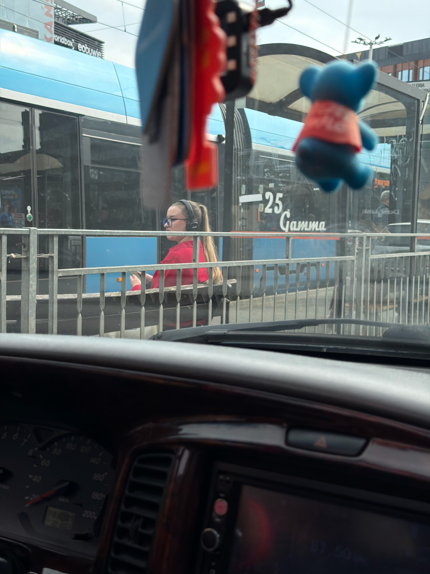 In Wrocław, Poland, a scene unfolds inside a vehicle, with the windshield partially obscured by dangling stuffed toys, a blue bear with an orange vest and a red object. Through the windshield, a tram is visible, bearing the number "25" and the word "Gamma." A young woman with blonde hair, wearing headphones and a red top, is seated inside the tram, holding a mobile phone. The interior of the vehicle features a wooden dashboard with a speedometer displaying readings up to 200 km/h and an illuminated display. The sky is overcast, suggesting a cloudy day.
