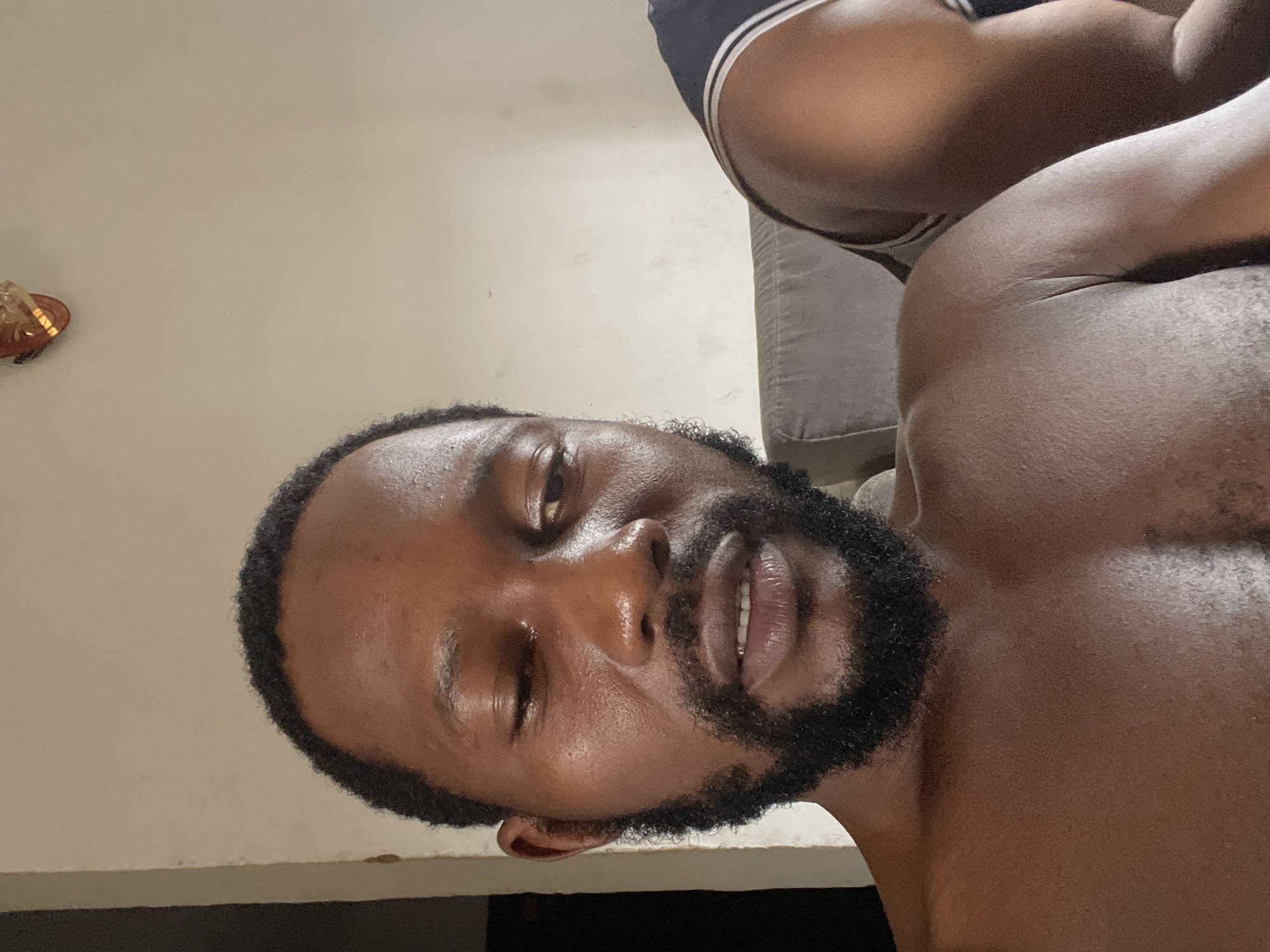 A shirtless man with a dark beard and short hair is pictured indoors. He is lying on a surface with a light-colored wall behind him. A small decorative item hangs on the wall in the upper left corner. To his right, another person's shoulder and part of their shirt are visible. There is a gray cushion to the right of the shirtless man. The location is likely a residential setting. Given the prompt's requirement, it is inferred that the location is in Abuja, Nigeria.