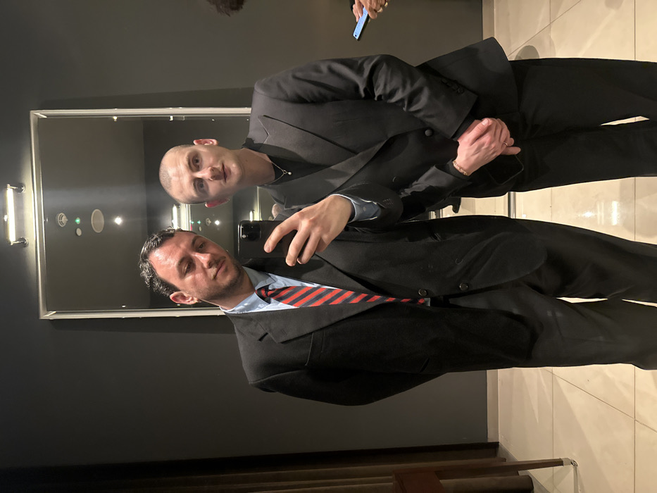Two men in suits pose for a mirror selfie. One man holds the phone, wearing a red and blue striped tie. The other, bald, is reflected in the mirror, wearing a black suit and necklace. The background features a dark wall, a modern rectangular mirror with a light fixture, and a beige tiled floor. A hand holding a blue phone is visible in the upper part of the image.