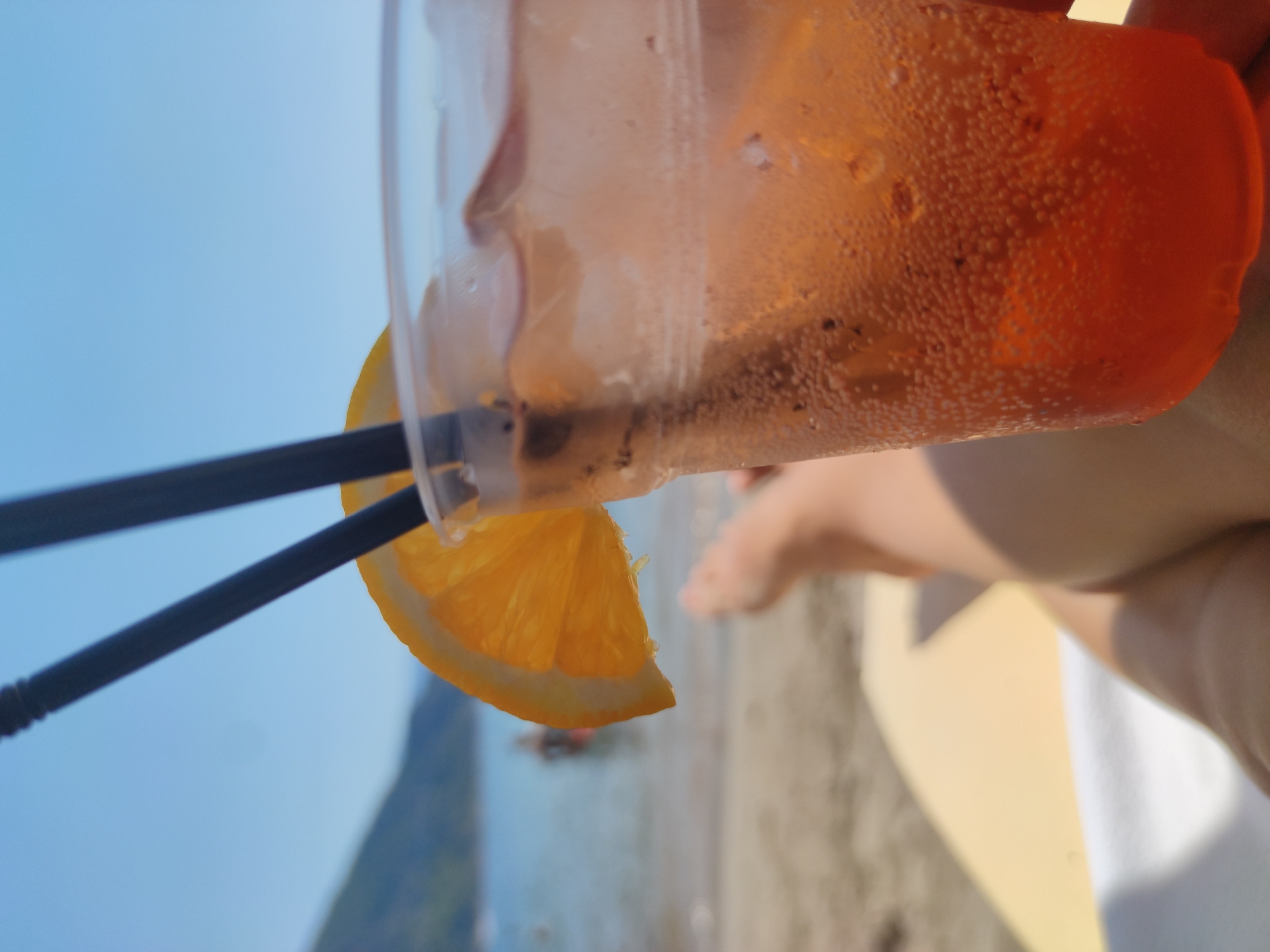 A close-up shot focuses on a cold drink in a clear plastic cup, garnished with a bright orange slice and two black straws. The drink itself is an amber color, filled with ice cubes and effervescent bubbles. In the background, a sandy beach stretches out, with the gentle blue waters of the sea and a distant, hazy shoreline. The sky above is a clear, bright blue. The lower portion of the image shows a person's legs resting on a yellow surface, suggesting they are lying down on a beach chair or lounger. The overall scene evokes a relaxed and tropical atmosphere.