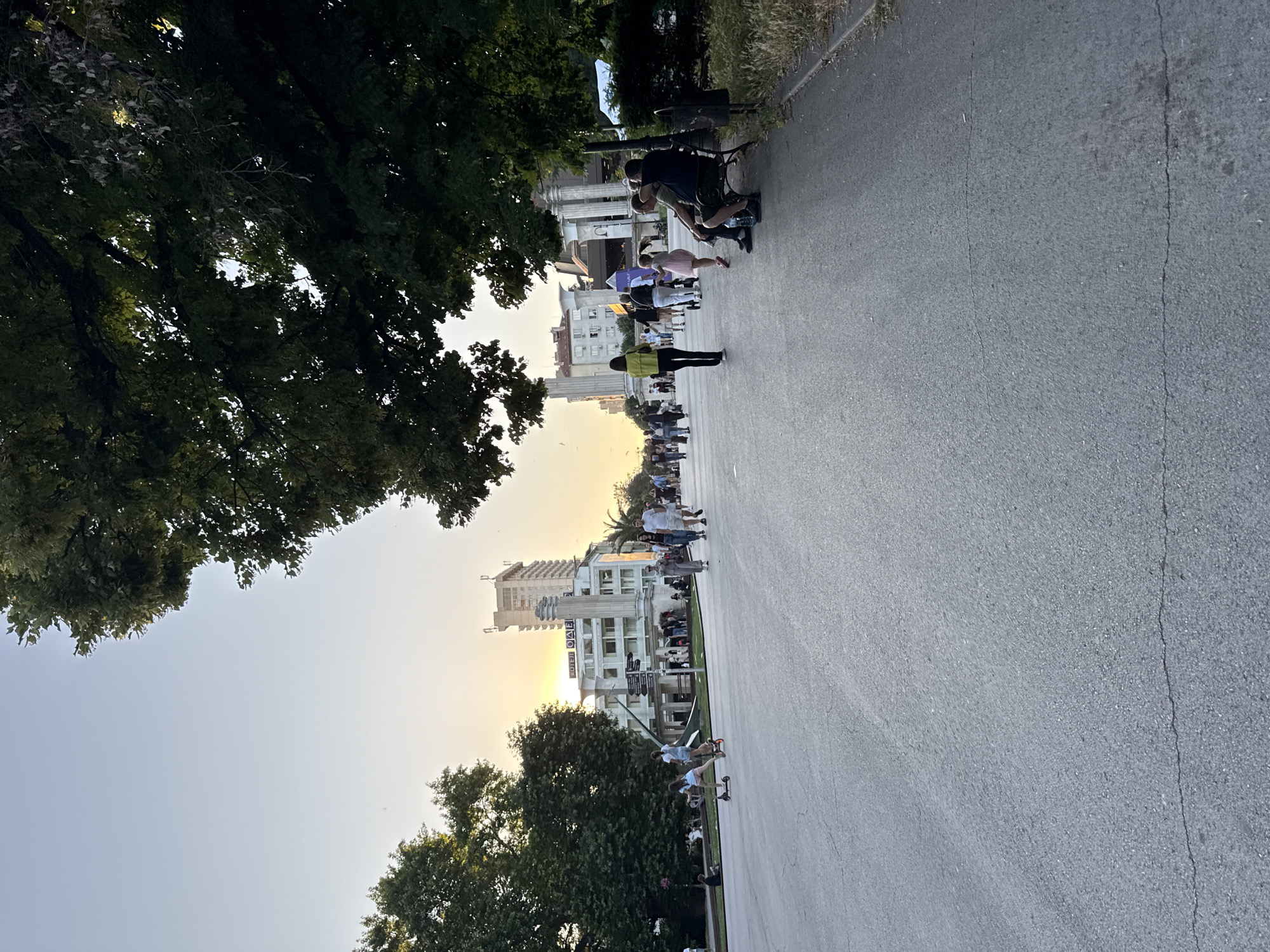 The image depicts a scene in a city park with a large paved area in the foreground. Several people are visible, walking, riding scooters, and sitting on a bench. In the background, there is a building with signs, and trees line the perimeter of the park. The sky is clear with a hint of sunset. The lighting suggests it is late afternoon or early evening.