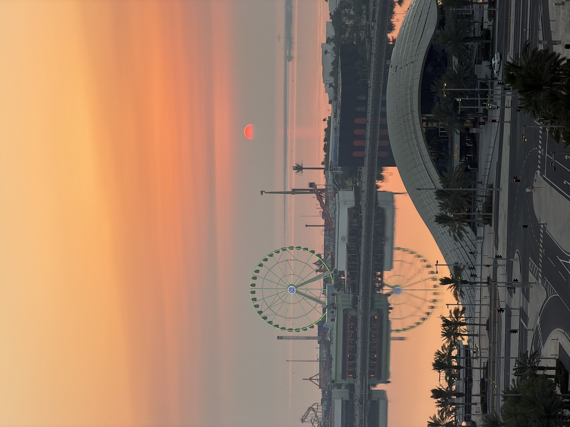 An aerial shot captures a waterfront amusement park at sunset in an unknown city. The sky is a gradient of orange and pink, with the sun, a small red circle, visible near the horizon. A large Ferris wheel, lit with green lights, stands prominently in the center of the park. Several other amusement park rides and structures are visible, including a roller coaster. In the foreground, a body of water reflects the Ferris wheel and the colorful sky. Beyond the water, a modern building with a distinctive curved roof and surrounding palm trees are seen, with a road and intersection with traffic lights in the immediate foreground. The scene appears to be a static, observational shot, with no people or animals clearly discernible.