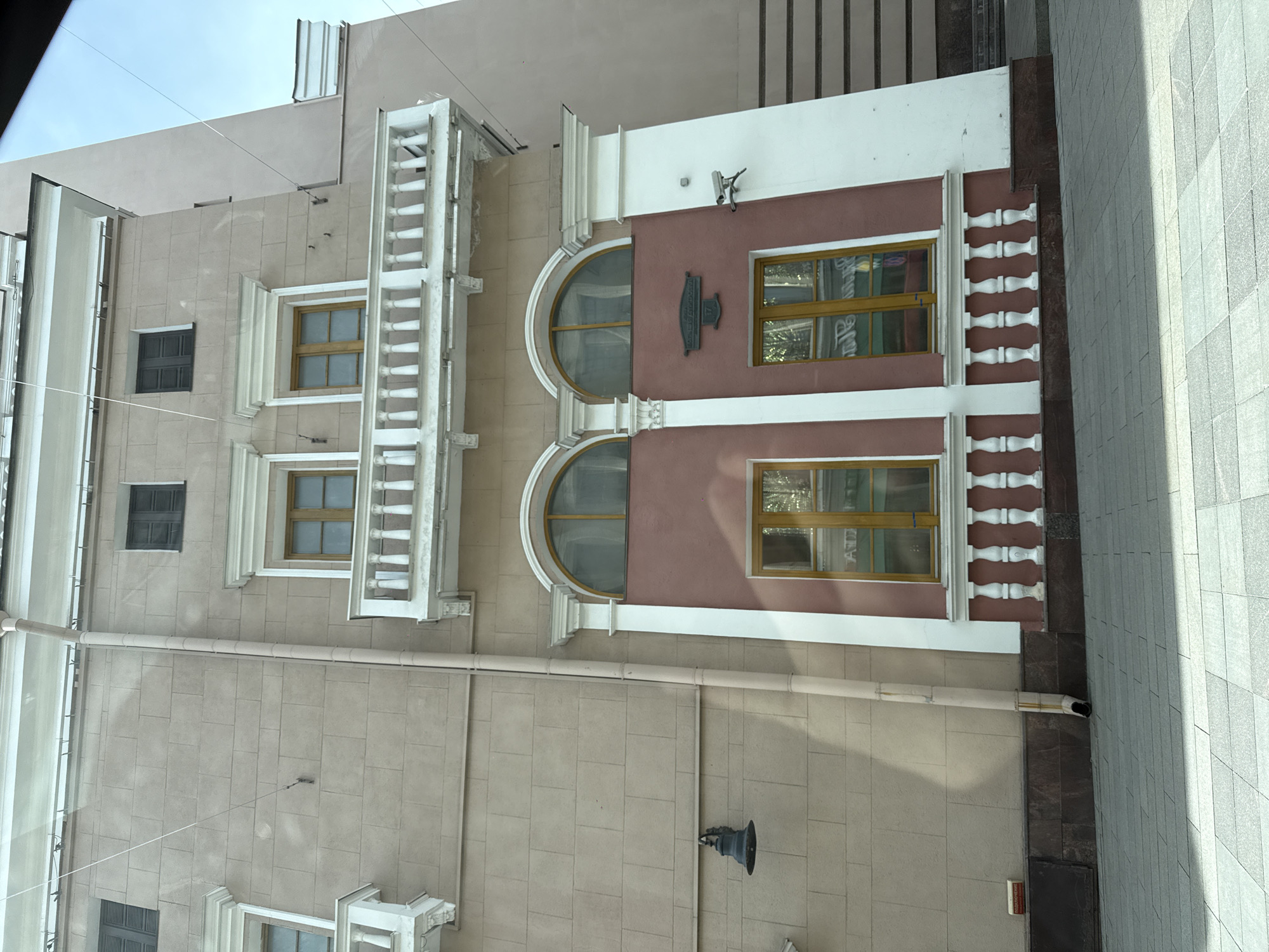 An architectural photograph captures a detailed section of a building facade in Moscow, Russia, seen from a slightly upward angle. The image is oriented vertically, showing the building's exterior with various architectural elements and windows.
The building features a multi-colored facade, primarily light beige or tan on the upper and lower sections, with a central reddish-brown portion. These sections are separated and adorned with white decorative moldings, cornices, and balustrades. On the left side of the upper beige section, there's a white balcony or ledge with a balustrade. Another, shorter white balustrade borders the lower part of the reddish-brown section on the right.
Windows are a prominent feature: two large arched windows are set within the reddish-brown section, framed by white molding. Above these, two rectangular windows with light-colored frames are visible on the beige wall. Further left, on the same beige wall, are two smaller rectangular windows fitted with dark grates or shutters. Reflections are visible in some of the windows, indicating the daytime setting.
A light-colored drainpipe runs horizontally across the beige facade before turning downwards. A black, ornate lamppost is affixed to the lower beige wall, and a small security camera is mounted on the white trim above the reddish-brown section. The building's base transitions into a darker, possibly granite or marble, material, bordered by a grey, tiled pavement on the right, suggesting a sidewalk.
A notable detail is a dark, rectangular plaque with an arched top, located above the upper arched window. It displays Cyrillic text, which reads "УЛ. СТАРАЯ БАСМАННАЯ" (Ul. Staraya Basmannaya), followed by the number "177". This indicates the building's address. The sky above the building appears clear and bright, consistent with a clear day. No people or overt activities are depicted, focusing solely on the building's structure and details.