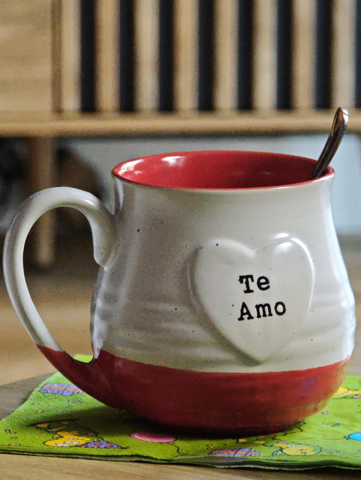 A white and red mug with the words "Te Amo" printed on a heart-shaped emblem is sitting on a green napkin with cartoon characters. A spoon is in the mug. The mug is on a wooden table. The background is blurred and shows vertical wooden slats. The location is not identifiable as Locking Parklands, United Kingdom.
