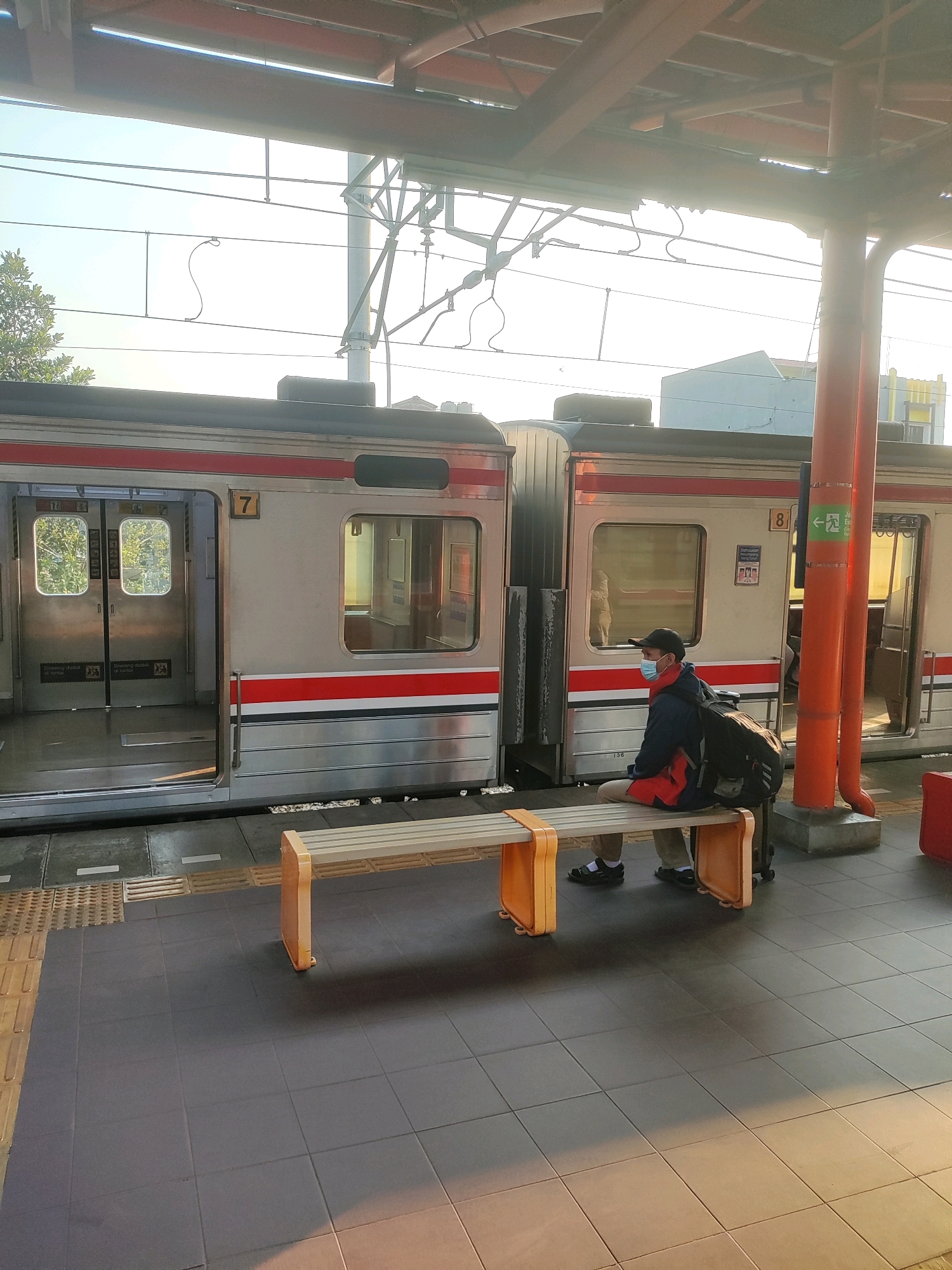 A person sits on a bench on a train platform in Bekasi, Indonesia, waiting for a train. The person is wearing a blue mask, a dark jacket with red accents, and khaki pants. They have a large backpack with them. The train has silver cars with red and blue stripes. The platform has a textured surface and a bench with orange supports. The overhead structure of the platform is made of metal beams and supports, with electrical wires visible above. The sun is casting long shadows across the platform, indicating it is likely early morning or late afternoon.
