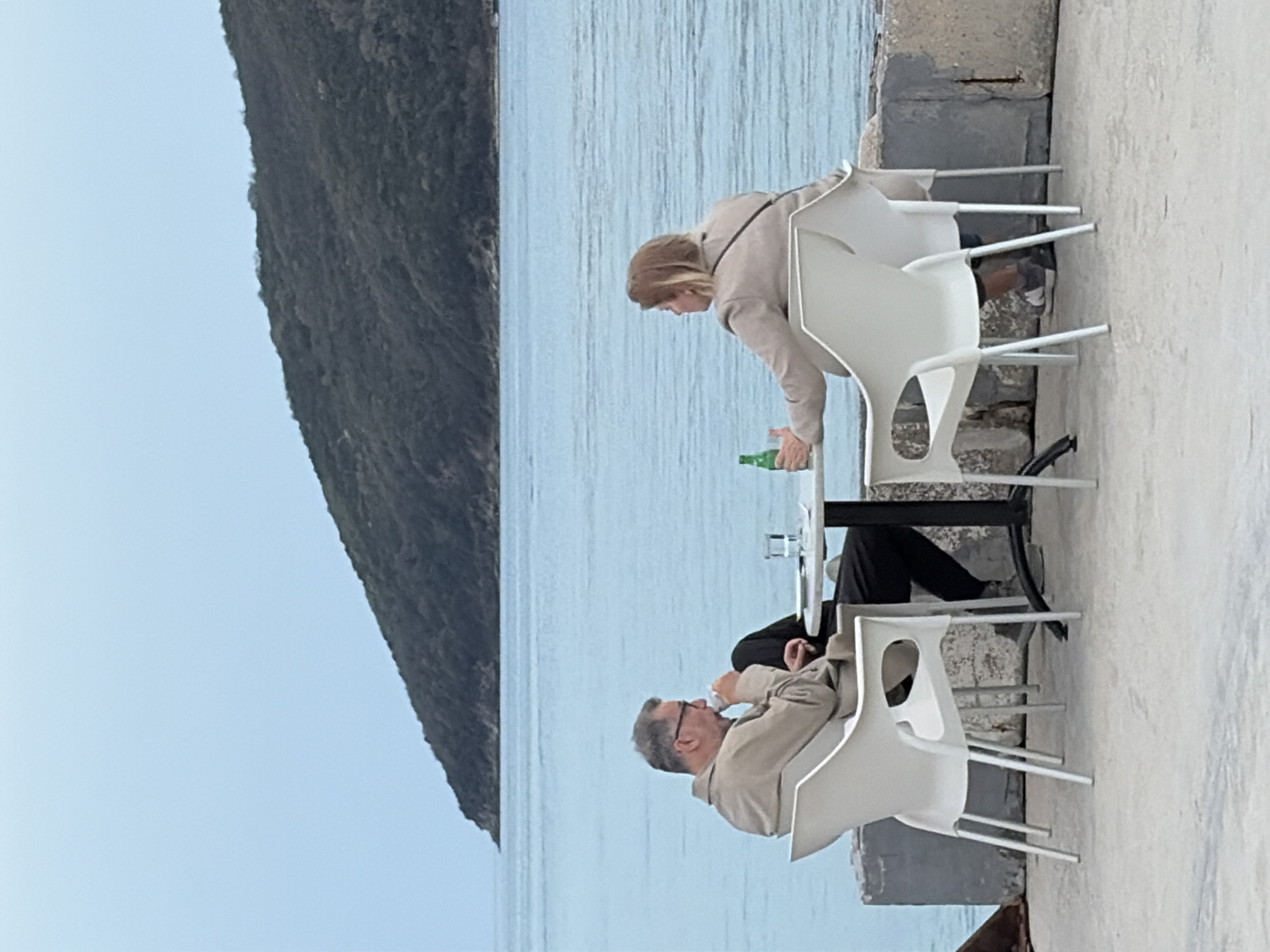 Two people sit at a small, round table on a concrete surface. The setting is near the sea, as suggested by the water and the large, tree-covered mountain visible in the background. A woman is reaching towards the table, possibly to place a green bottle down. A man sits opposite her, drinking from a clear glass. Both are wearing light-colored clothing. The table is black and stands on a light concrete surface near a retaining wall. The scene appears to be an outdoor dining area, possibly in Budva, Montenegro, given the typical Adriatic coastline scenery. The chairs are white plastic or resin and are similar in design.