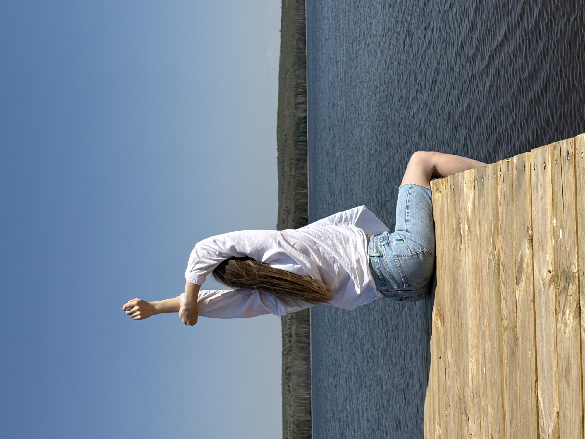 A photograph taken in Chaikovsky, Russia depicts a person sitting on the edge of a wooden dock or pier. The person is wearing jean shorts and a white shirt and is leaning forward, extending their arms outward. The body of water is behind the dock, with a shoreline visible in the distance. The sky is clear and blue.