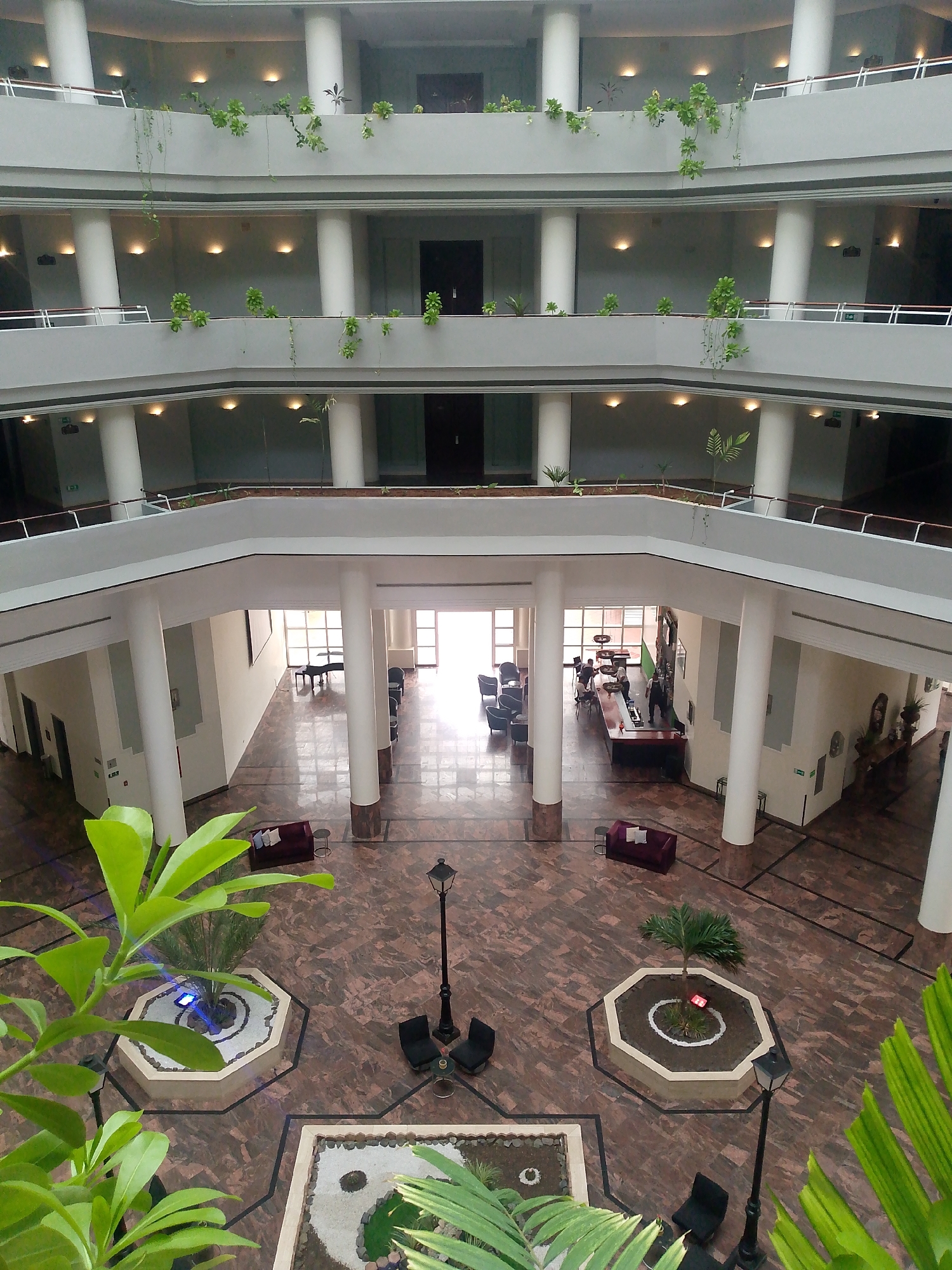The image depicts the interior atrium of a multi-story building, possibly a hotel or office complex. The atrium features a central open space with marble floors and a polished dark red and brown patterned tile. Several levels of balconies overlook the central area, each lined with white cylindrical columns and potted plants. The upper levels are illuminated by recessed ceiling lights.
On the ground floor, the atrium has a bar area with patrons and staff. A grand piano is visible in a waiting area near large windows that allow natural light to enter. In the central open space, there are decorative planters with small trees and plants, as well as street lamps and seating arrangements. Two maroon sofas are placed on the floor.
The image also shows foreground foliage from plants, suggesting that the photo was taken from a higher vantage point within the building, looking downwards into the atrium. The overall impression is one of a modern, well-maintained indoor public space. There is no visible signage or context to definitively place this location in Pedro González, Venezuela, or any other specific city.
