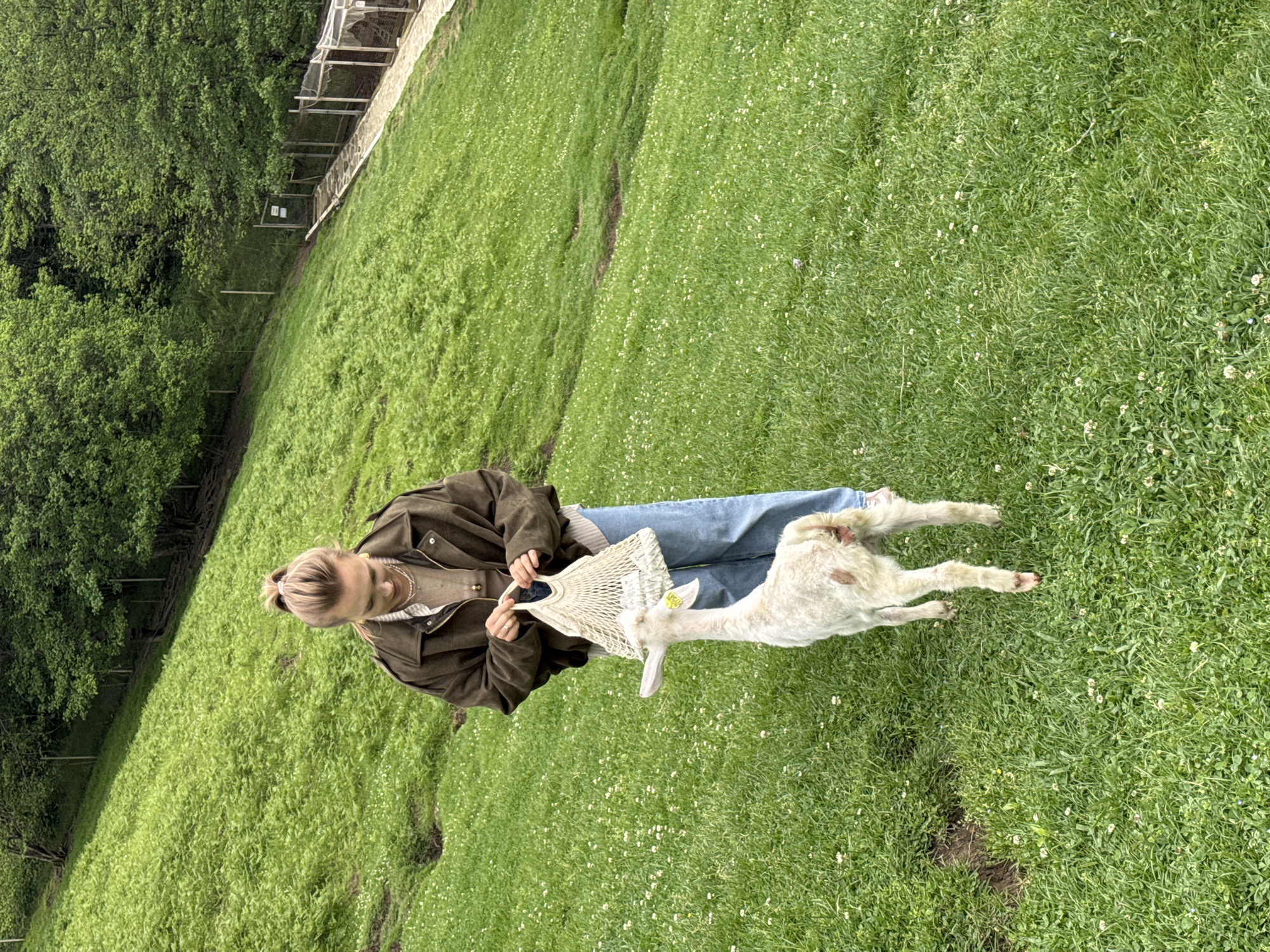A woman in a green jacket and blue jeans stands on a grassy field, holding a white mesh bag. A small white goat with brown spots is eating from the bag. The field is covered in green grass and small white flowers. In the background, a fence borders the field and trees are visible. The location is implied to be in the city of Progress, Russia.