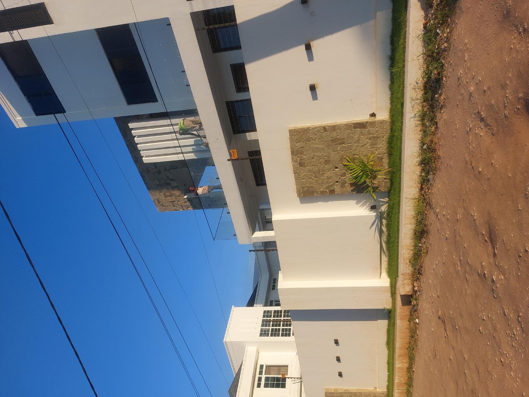 The image depicts a modern white building in Dar es-Salaam, Tanzania, under a clear blue sky. Two individuals are visible on the building's glass-covered balcony, one wearing a green vest. The building features large windows and stone accents on the façade. A small palm tree grows near the building's base along the edge of a dirt road. Power lines intersect the sky above the building.