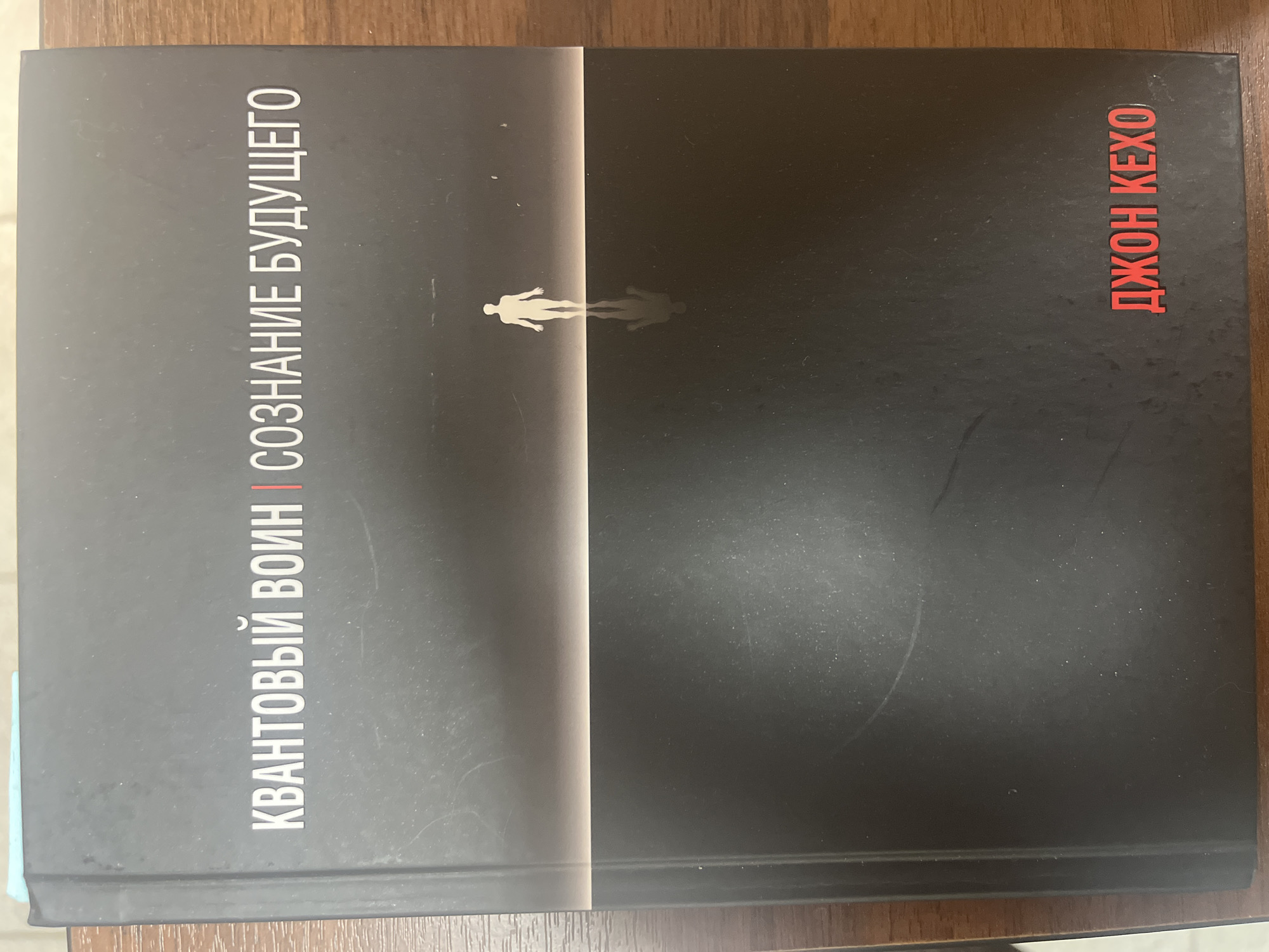 The image shows a book lying on a wooden surface. The book cover is primarily black with a vertical white stripe separating the left and right sides. A white silhouette of a person and their shadow is visible on the stripe. The title "КВАНТОВЫЙ ВОИН | СОЗНАНИЕ БУДУЩЕГО" is printed vertically in white on the left side. The author's name, "ДЖОН КЕХО," is printed vertically in red on the right side. The photo was taken indoors and no information indicates the location is in Ulyanovsk, Russia.