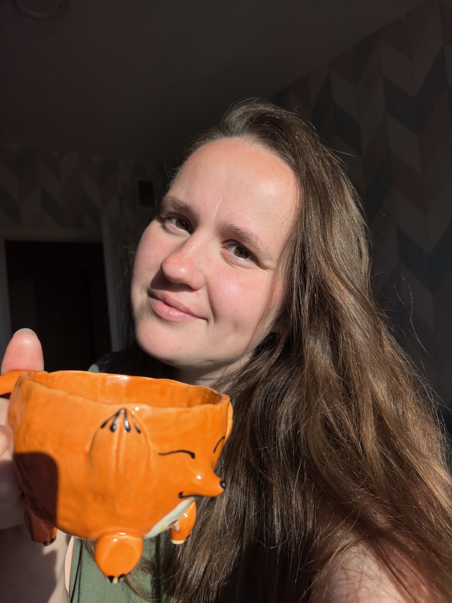 A medium shot shows a woman with long, brown hair and fair skin holding an orange ceramic mug shaped like a fox. The mug has black accents for eyes, nose, and paws. The woman is smiling and looking at the camera. Sunlight is casting a shadow across her face from the left side of the frame. The background is out of focus, showing a wall with a geometric pattern and a dark area that could be a window or doorway. The scene appears to be indoors, possibly in a residential setting in Фролы, Russia.