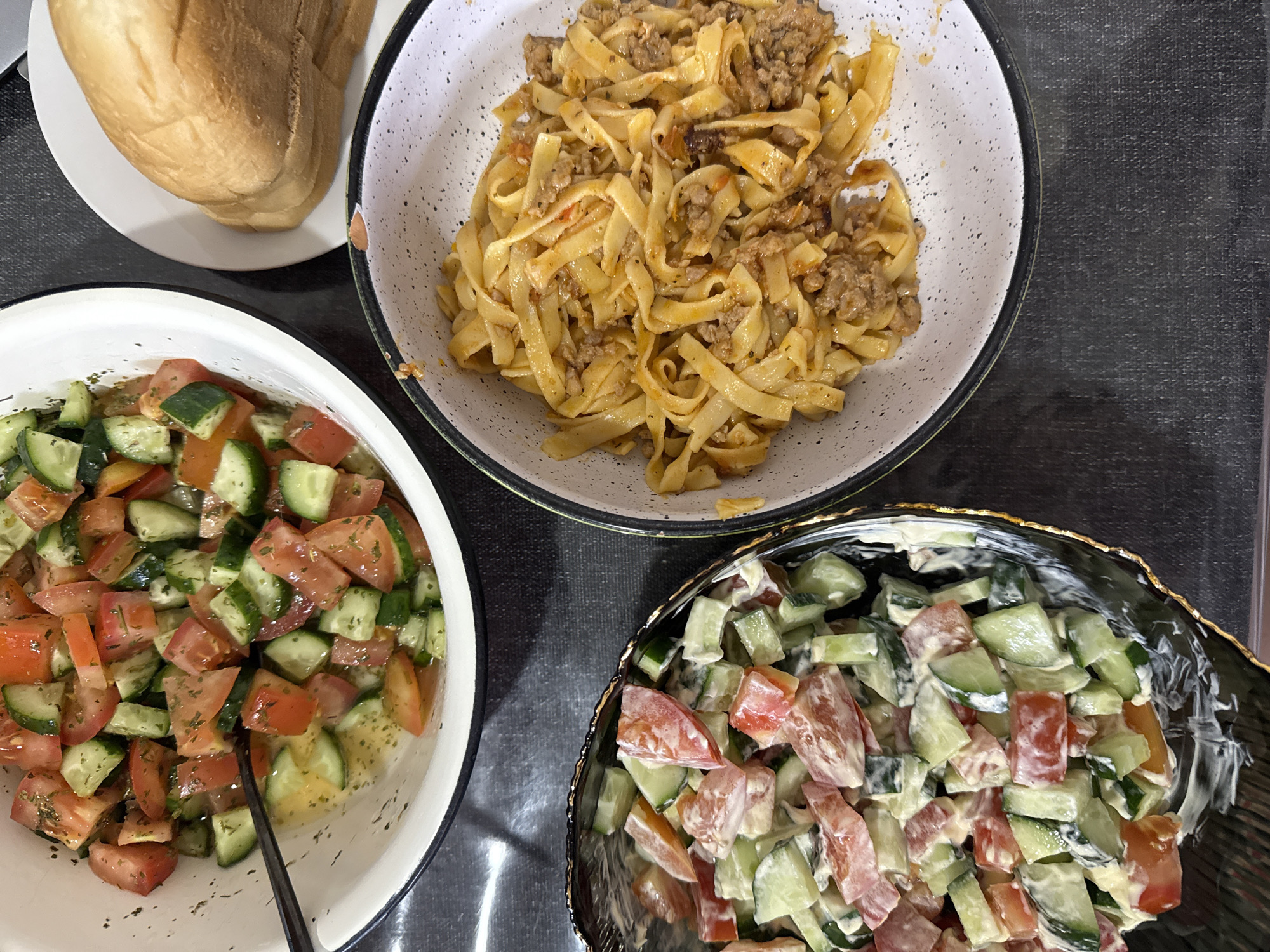 The image shows a table with three bowls and a plate, suggestive of a meal setting. On the top left, there is a plate with what appears to be bread. To the right of the bread, there's a bowl of tagliatelle pasta with meat sauce. Below the bread and pasta is a bowl containing a salad of tomatoes and cucumbers with a light dressing. To the right of the salad is a bowl with a similar salad, but with a creamy dressing. The background is a dark surface with a visible texture. The specific location, Khimki, Russia, is not evident from the image, as there are no distinguishing landmarks or contextual clues within the image.