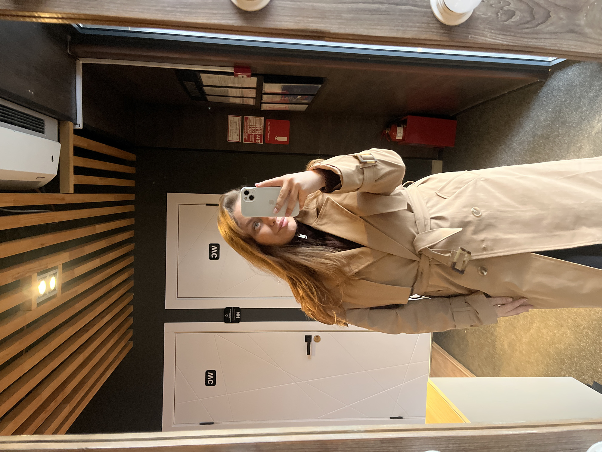 A young woman with long, light brown hair, wearing a beige trench coat over a dark top, is captured in a mirror selfie. She holds a light-colored smartphone (likely an iPhone) to take the photo, partially obscuring her face as she looks into the phone. The setting, reflected in a large mirror, appears to be an interior public space, potentially a hallway or common area in Chaikovsky, Russia.
To the left of the woman in the reflection, a wall features horizontal wooden slats and a light fixture with two bright bulbs. A white air conditioning unit is mounted high on the wall in the upper left corner. To her right, two white doors with a subtle geometric pattern are visible. Each door has a black handle and a black square sign with "WC" written in white, appearing upside down due to the mirror reflection. Between the doors, a universal accessibility symbol is present. Above the doors, on a dark wall, are several framed notices or signs, including a red one with Cyrillic text, partially readable as "ОБЯЗАТЕЛЬНО" (meaning "obligatory" or "required"). A red fire extinguisher is also mounted on the wall further to the right. The floor has a textured, grey, possibly concrete-like appearance. The bright artificial lighting suggests an indoor environment. The woman's expression is neutral, focusing on her phone as she takes the picture.