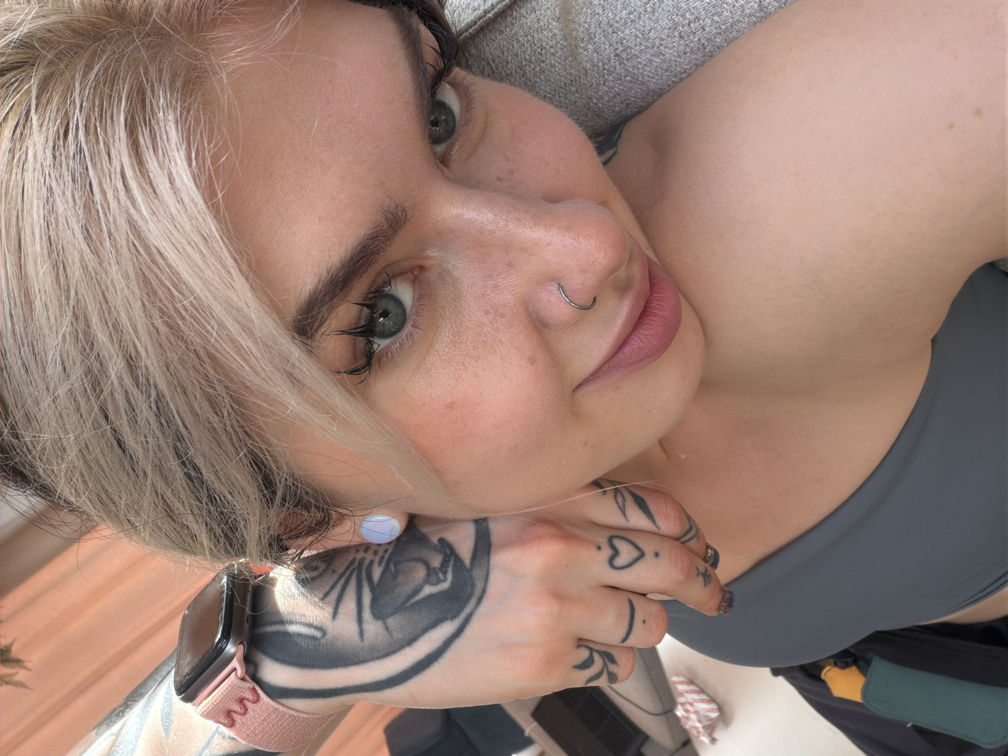 A selfie-style, medium shot depicts a fair-skinned individual with ash-blonde hair and green eyes. She is looking at the camera with a slight smile, sporting a nose ring. She is wearing a gray sports bra. Prominent tattoos are visible on her left hand and forearm, including a large design on the forearm and smaller symbols on her fingers. A smartwatch with a pink band is worn on her left wrist. The background is soft and suggests an indoor setting. There is no information to infer the location in the city of Budva, Montenegro.