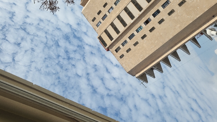 The scene presents an upward-angled view of urban architecture against a sky in Magaluf, Spain. The primary subject is a multi-story building, oriented diagonally from the bottom-right towards the upper-left. Its facade is constructed of light tan masonry units, featuring multiple rectangular windows and a distinctive series of triangular projections or balconies along its left-facing side. Horizontal dark-slatted vents are integrated into upper floor sections, and a segment of a red rooftop is visible at its apex. A second, lighter-toned building is partially visible along the extreme left edge of the frame, showing a vertical segment of its facade with a lighter trim. The sky above is light blue, extensively covered with patchy, white altocumulus-like clouds. Fine, dark linear elements, consistent with utility wires, extend diagonally across the mid-ground. Bare tree branches are visible against the sky in the upper-right corner. A white, cylindrical object with a rounded cap is partially present at the bottom-right.