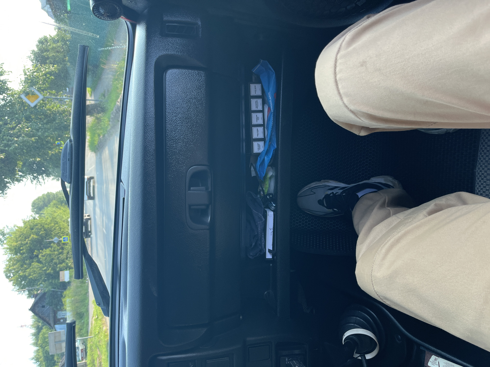 The image shows the interior of a car, looking forward through the windshield. The driver's legs, clad in light tan pants and athletic shoes, are visible in the foreground. To the left, a gear shift knob is partially visible. In the center of the dashboard, the glove compartment is open, revealing several small, rectangular boxes of what appear to be perfumes, labeled "YVES SAINT LAURENT," "TOM FORD," "CHRISTIAN DIOR," "ESCENTRIC MOLECULES," and "HUGO BOSS." A blue package, possibly containing wipes, is also in the glove compartment. Outside the car, a paved road can be seen, with trees and a wooden house lining the sides. A yellow diamond-shaped road sign with a black border is visible in the distance. The scene suggests a drive through a rural area in or near Chaikovsky, Russia, given the presence of Russian text on the blue package and the architectural style of the house. The lighting indicates it is daytime.
