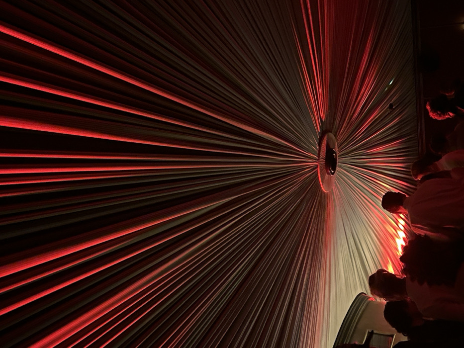 A dimly lit space features a wall installation that resembles a starburst pattern. Numerous lines emanate from a central disc, creating a radial effect. The lines are illuminated with red light, creating a contrast against the darker background. People are visible in the lower right, suggesting the space is occupied by attendees. The perspective is from a low angle, enhancing the sense of depth and scale.