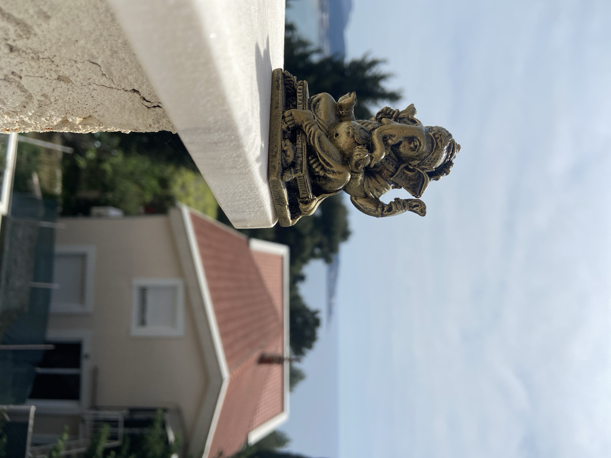 A bronze-colored statue of Ganesha sits on a white marble ledge. In the background is a house with a red tile roof, trees, and a body of water under a light blue sky. The location is likely Šušanj, Montenegro.