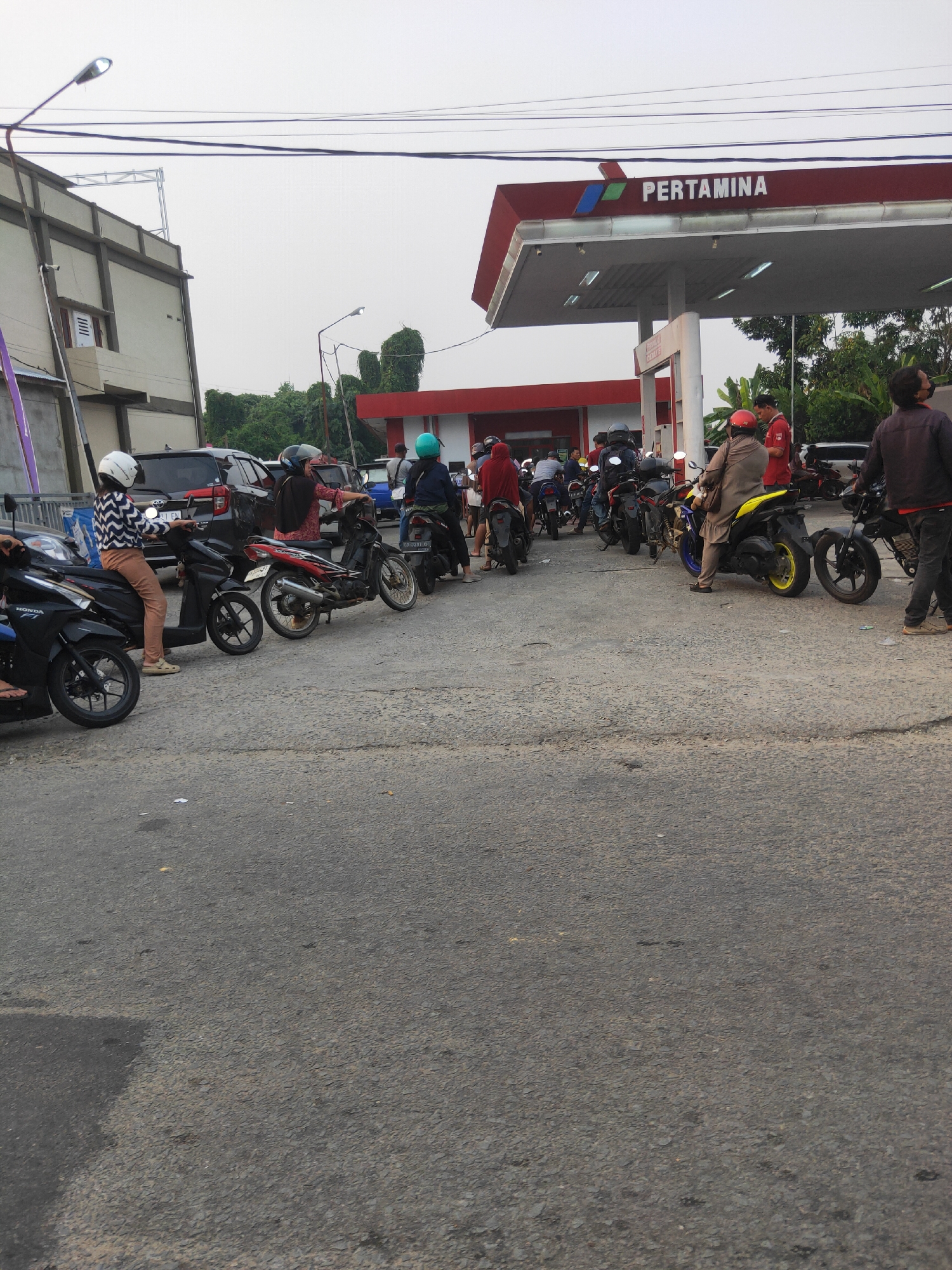 This image depicts a queue of motorcycles and their riders at a Pertamina gas station in Sintang, Indonesia. The gas station canopy is red with the Pertamina logo prominently displayed. Several individuals are on or near their motorcycles, some appearing to be waiting for service. The ground is paved asphalt, and a building with a modern facade is visible to the left of the gas station. The overall scene suggests a typical day at a busy gas station.