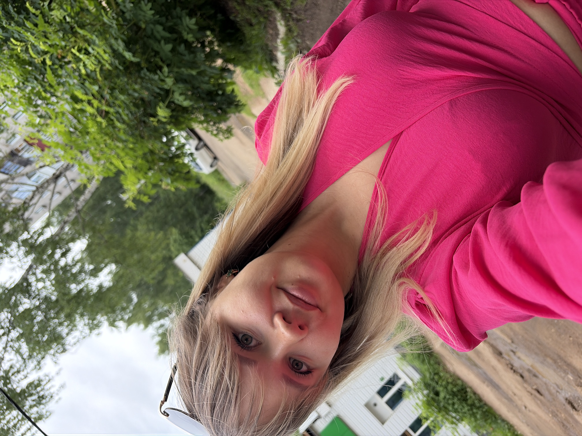 A young woman with blonde hair and green eyes, wearing a bright pink V-neck top and sunglasses, poses for a selfie outdoors. She is positioned slightly off-center in the frame, with her face tilted towards the camera and a slight smile. Her blonde hair is worn down, with bangs framing her face. The background suggests an urban environment, with a building visible on the left, possibly a multi-story apartment block, and lush green trees and foliage surrounding the scene. A dirt road with some puddles is visible in the foreground, and a white car is parked on the road in the distance. The sky is overcast. The image appears to be taken in Chaikovsky, Russia, given the presence of typical residential buildings and natural elements found in the city.