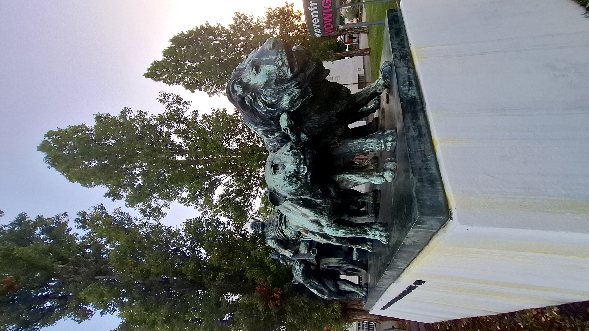 A bronze sculpture of a Roman-style chariot pulled by four lions, with a man in Roman attire driving, is positioned on a white stone pedestal. The sculpture, weathered to a verdigris patina, depicts the lions in motion, their bodies tense and muscles defined. The man sits upright in the chariot, his gaze directed forward. The chariot has two spoked wheels visible on the left side. The pedestal exhibits streaks of yellow discoloration running down its white surface, suggesting water runoff. In the background, tall, slender trees with dense green foliage reach towards a pale sky. To the right, a building is partially visible, along with a sign featuring pink lettering that reads "horenfr LUDWIG". The overall setting appears to be a public space, likely a park or square. The sculpture is located in front of the Beethoven-Gedenkstätte (Beethoven Memorial) in Heiligenstadt, Vienna, Austria.