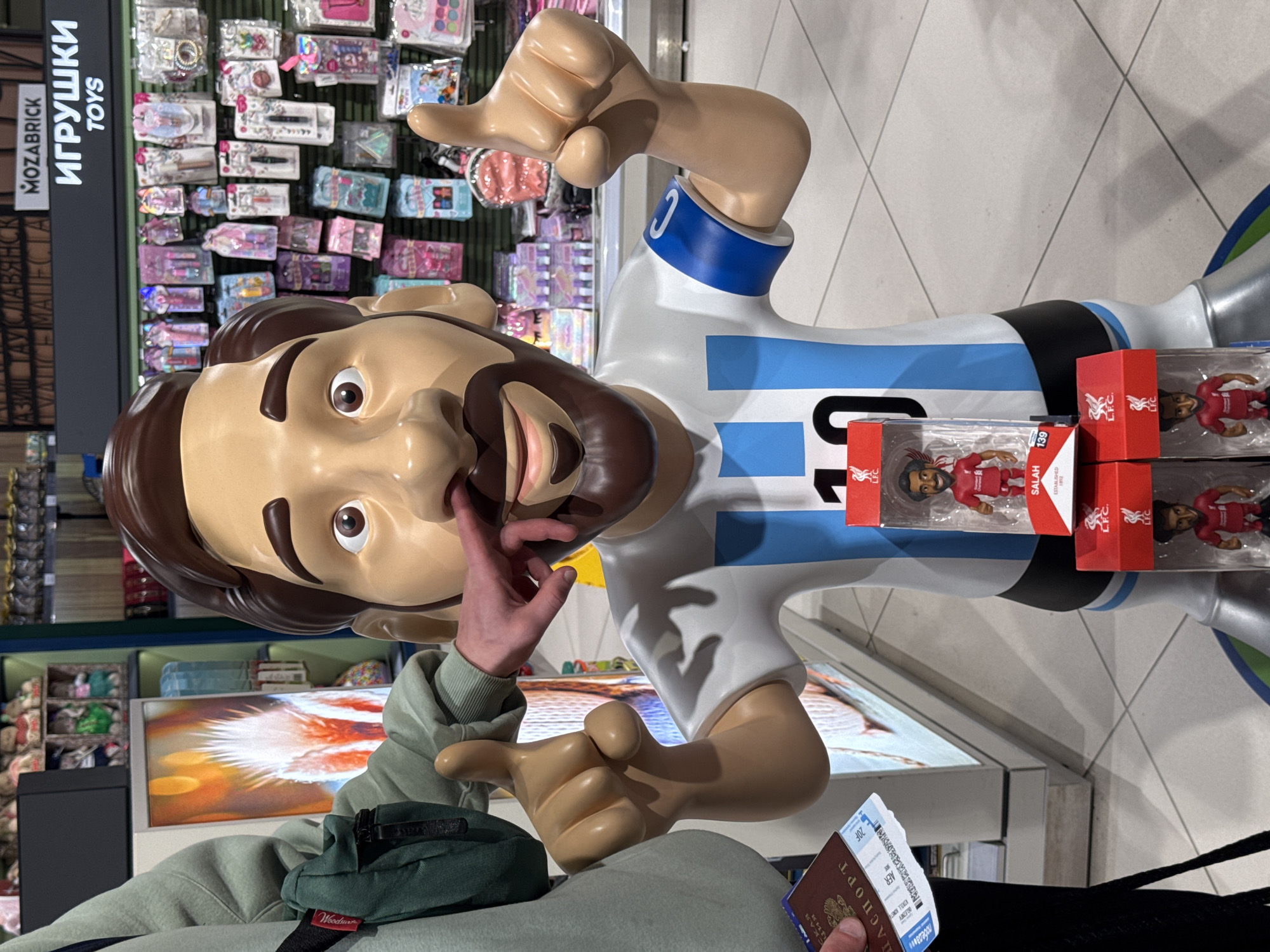 The image shows a tourist posing with a statue of Lionel Messi, wearing the Argentina national football team jersey, inside what appears to be a shop. The tourist is holding a passport and plane ticket in one hand and touching the statue's face with the other. Next to the statue, there are boxes of Liverpool Football Club (LFC) Mohamed Salah figures on display. The shop is stocked with various toys, and signage with the words "ИГРУШКИ TOYS" (meaning "Toys" in Russian) and "MOZABRICK" is visible. The text on the passport is in Russian, and the plane ticket mentions the city of Adler/Sochi.