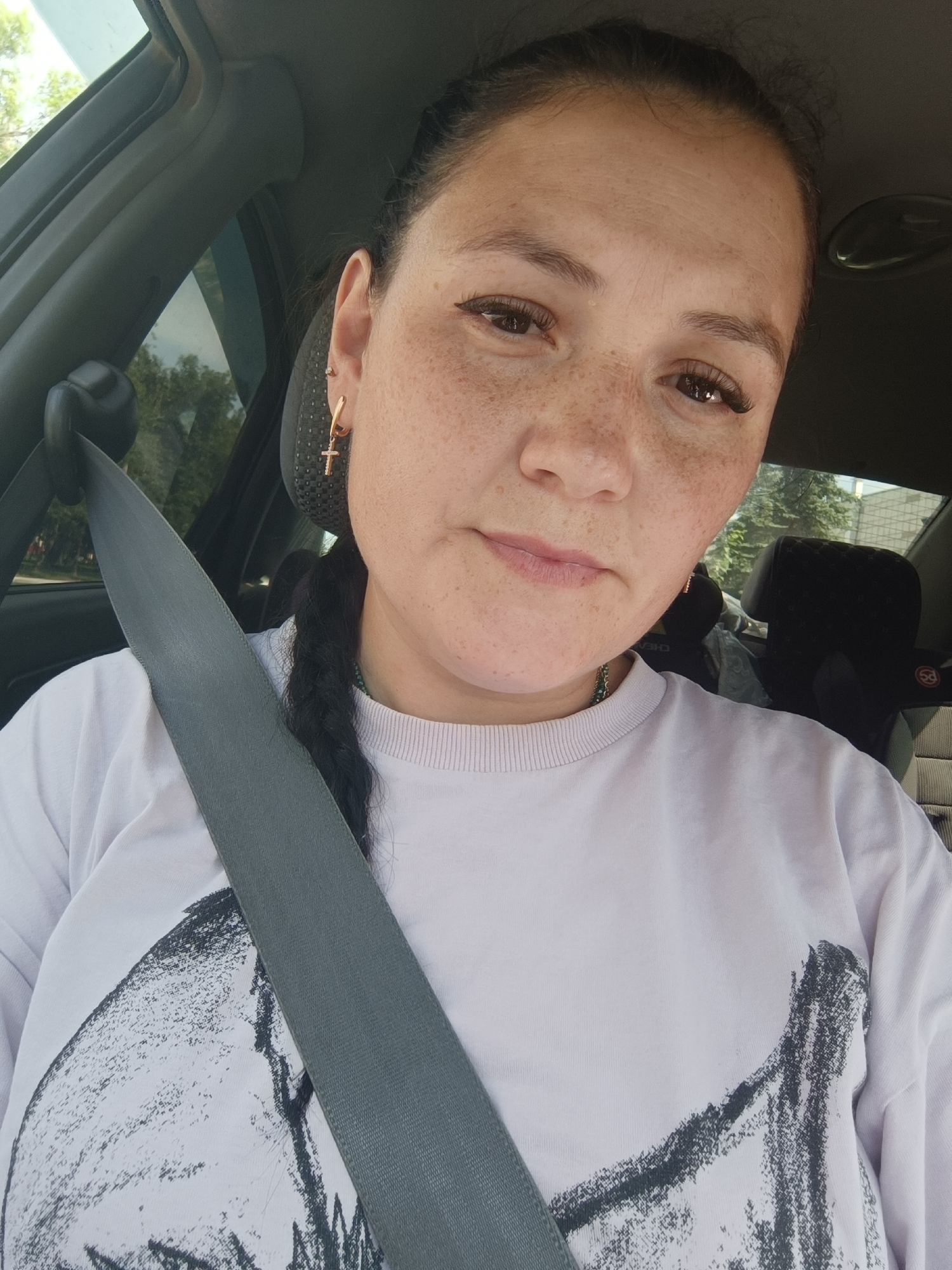 A woman is in the driver's seat of a car, wearing a light-colored t-shirt and a seatbelt. She has dark hair pulled back in a braid and is wearing a gold cross earring. She has a light complexion with freckles across her nose and cheeks. The interior of the car is visible behind her, including a headrest and a portion of the passenger seat. The background outside the car windows shows trees and foliage, suggesting a daytime scene. The image is taken from a selfie perspective.