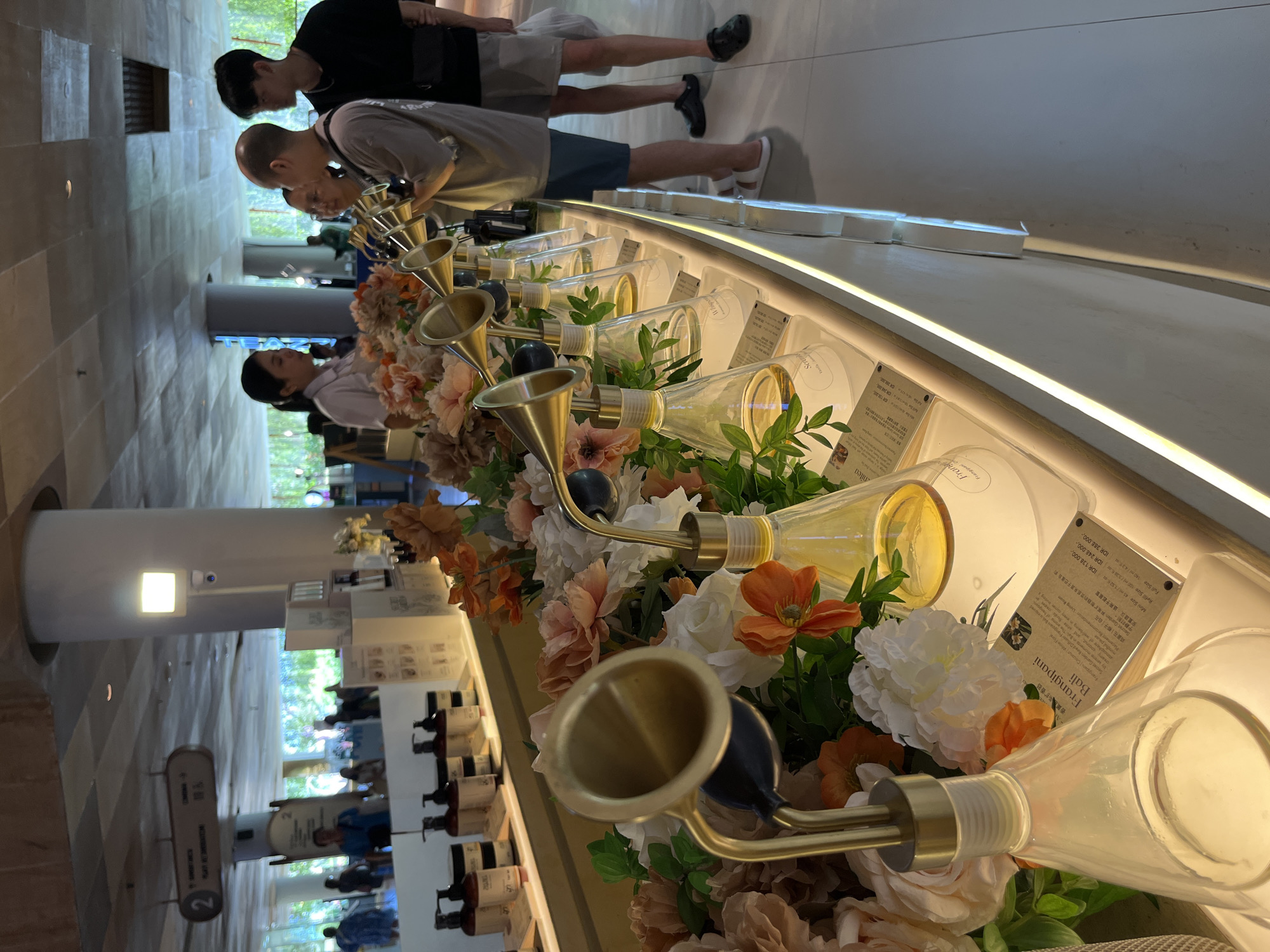 The image depicts a group of people interacting with a display of fragrance diffusers in what appears to be a retail or boutique setting. The subjects are primarily young adults, with several individuals leaning in to smell the products. The diffusers are housed in clear glass bottles with decorative elements, including faux flowers and metallic spouts, suggesting a focus on sensory experience and aesthetic appeal.
The scene is indoors, with soft lighting that creates a warm and inviting atmosphere. The background suggests a modern interior with tiled walls and flooring, and glimpses of greenery and other store elements. One of the individuals is wearing a white shirt, possibly an employee or uniform.
Prominently displayed is text from a product label, which reads "Frangipani Bali," along with details about the fragrance notes ("dreamy coconut, and sh," "鸡蛋花椰子|白花") and placement recommendations ("Living Room"). Pricing information in Indonesian Rupiah (IDR) is also visible, indicating that the location is likely in Indonesia, as specified by the context "Kuta, Indonesia".
The overall impression is one of a leisurely shopping experience, with customers engaging with the products in a calm and appreciative manner. The floral decorations and the nature of the products suggest a focus on relaxation and well-being. The presence of a sign with the number "2" could indicate a specific section or department within the establishment. The time of day is not explicitly discernible, but the indoor lighting implies it could be any time.